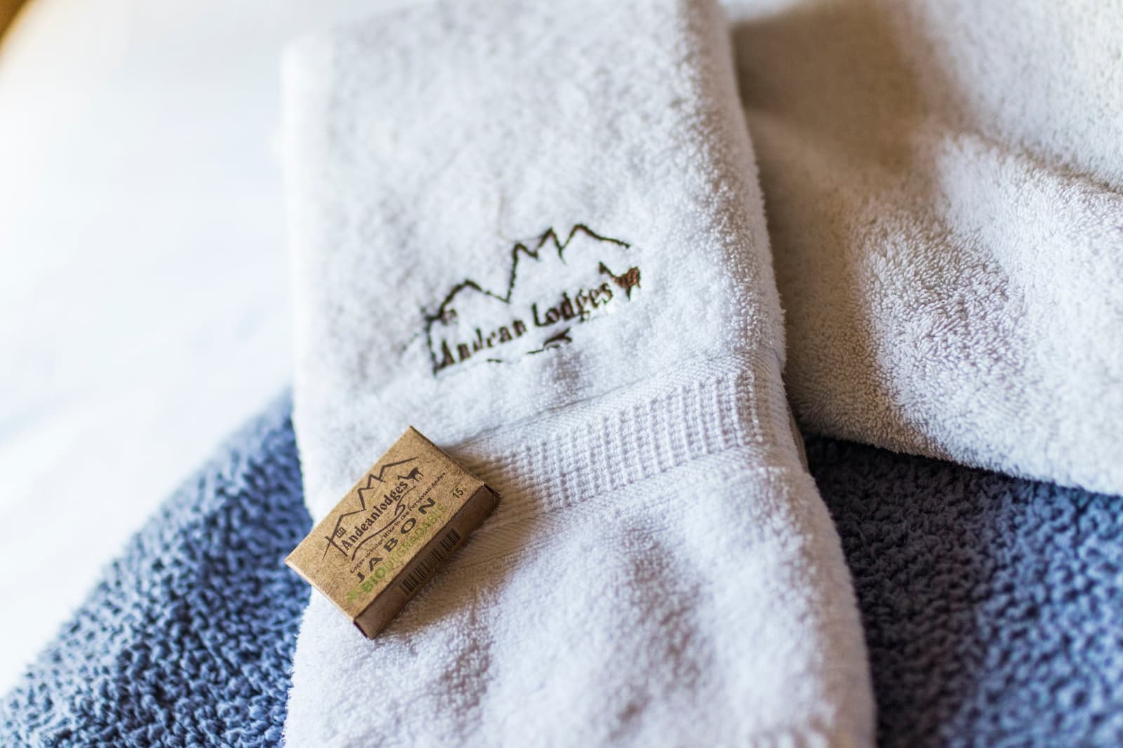 Biodegradable soap, soft towels, hot water. Small details that matter after a long day above 4,800 m. Photo: Diego Nishiyama - Fotografia White fluffy towel embroidered with the Andean Lodges logo folded on a blue towel, with a small biodegradable soap bar in kraft packaging resting on top, part of the Ausangate Lodge Trek accommodation experience