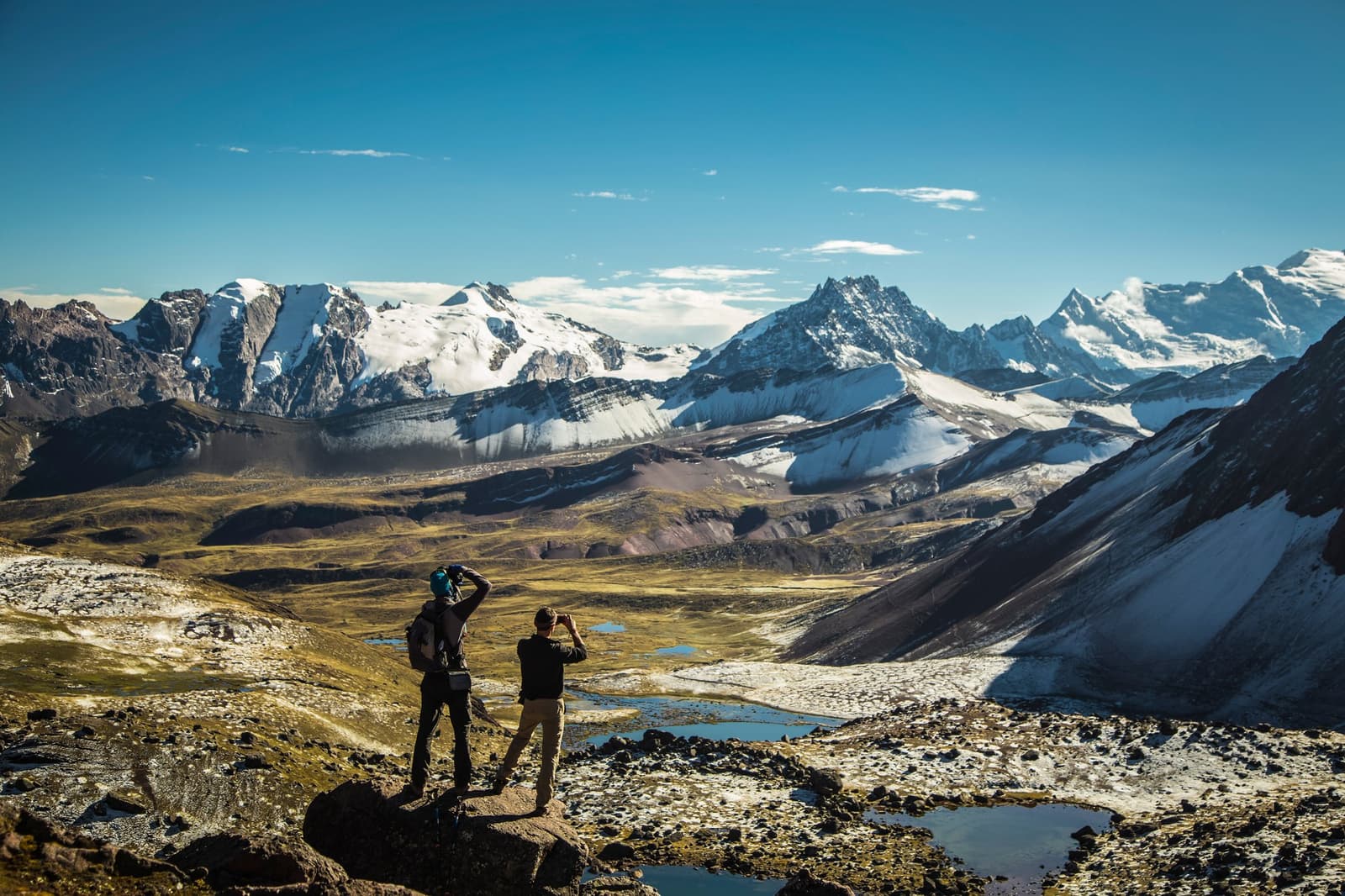 Five seconds at the top, binoculars out, trying to take it all in at once. Photo: Declercq Two hikers stand on a rocky ridge on the Ausangate Lodge Trek, scanning a wide high-altitude valley with scattered glacial pools, puna grassland, and a panorama of snow-capped Andean peaks under a deep blue sky