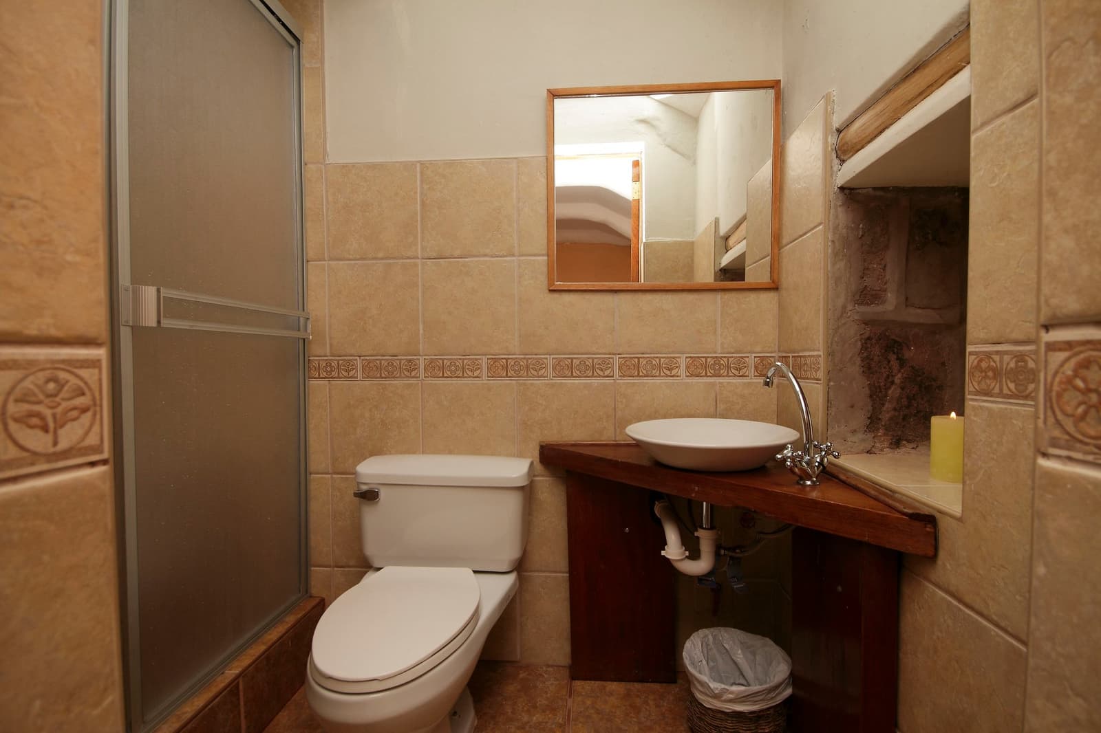 Hot water at 4,800 m. You've earned it. Photo: Gustavo Vivanco Leon Private en-suite bathroom in an Ausangate Lodge Trek stone cabin, featuring a vessel sink on a wooden vanity, toilet, frosted-glass shower cubicle, decorative ceramic tile border, and a lit candle on the shelf