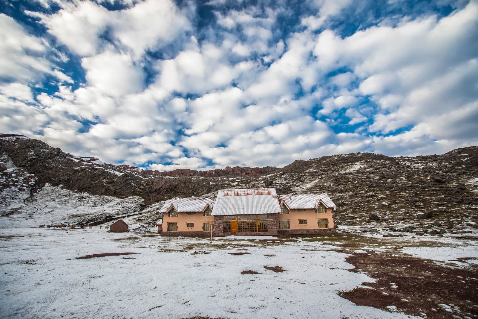 Snow arrived overnight. The lodge didn't mind, and neither should you. Photo: CH DECLERCQ A stone and plaster mountain lodge dusted with snow sits alone in a high-altitude valley on the Ausangate Lodge Trek, surrounded by rocky peaks under a wide, cloud-filled sky