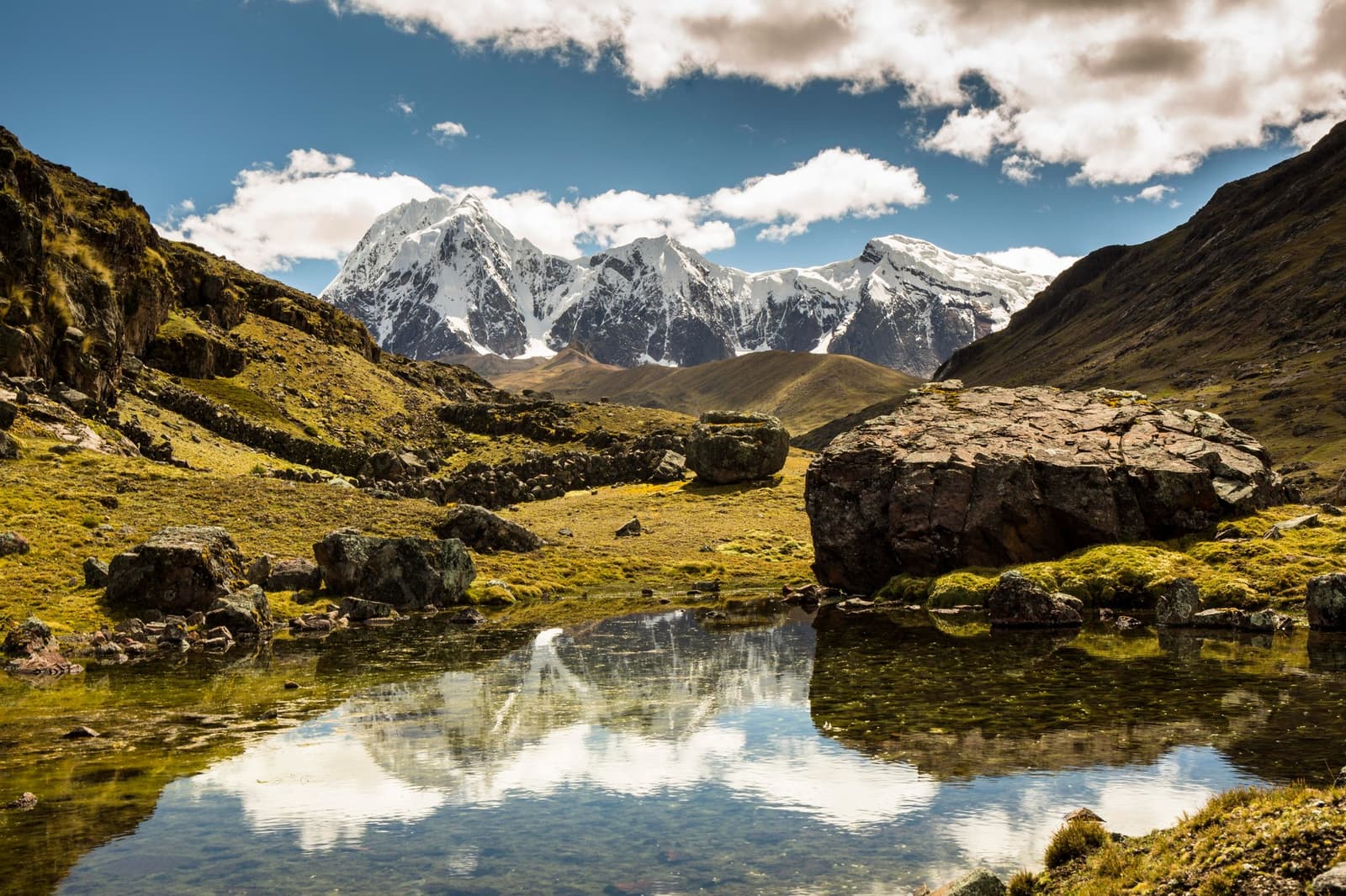 Cold, clear, and completely unbothered by the altitude. A still alpine tarn in a high-altitude Andean valley reflects snow-capped peaks and clouds during the ausangate trek, with moss-covered boulders and yellow-green puna grassland stretching toward the mountains