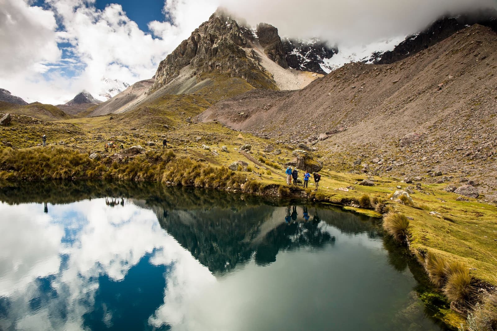 The lake so still it doubles the mountain. Stop here as long as you need. A small group of hikers on the ausangate trek pauses beside a still high-altitude glacial lake reflecting rocky snow-dusted peaks and clouds, with ichu grass on the banks and pack animals visible in the distance