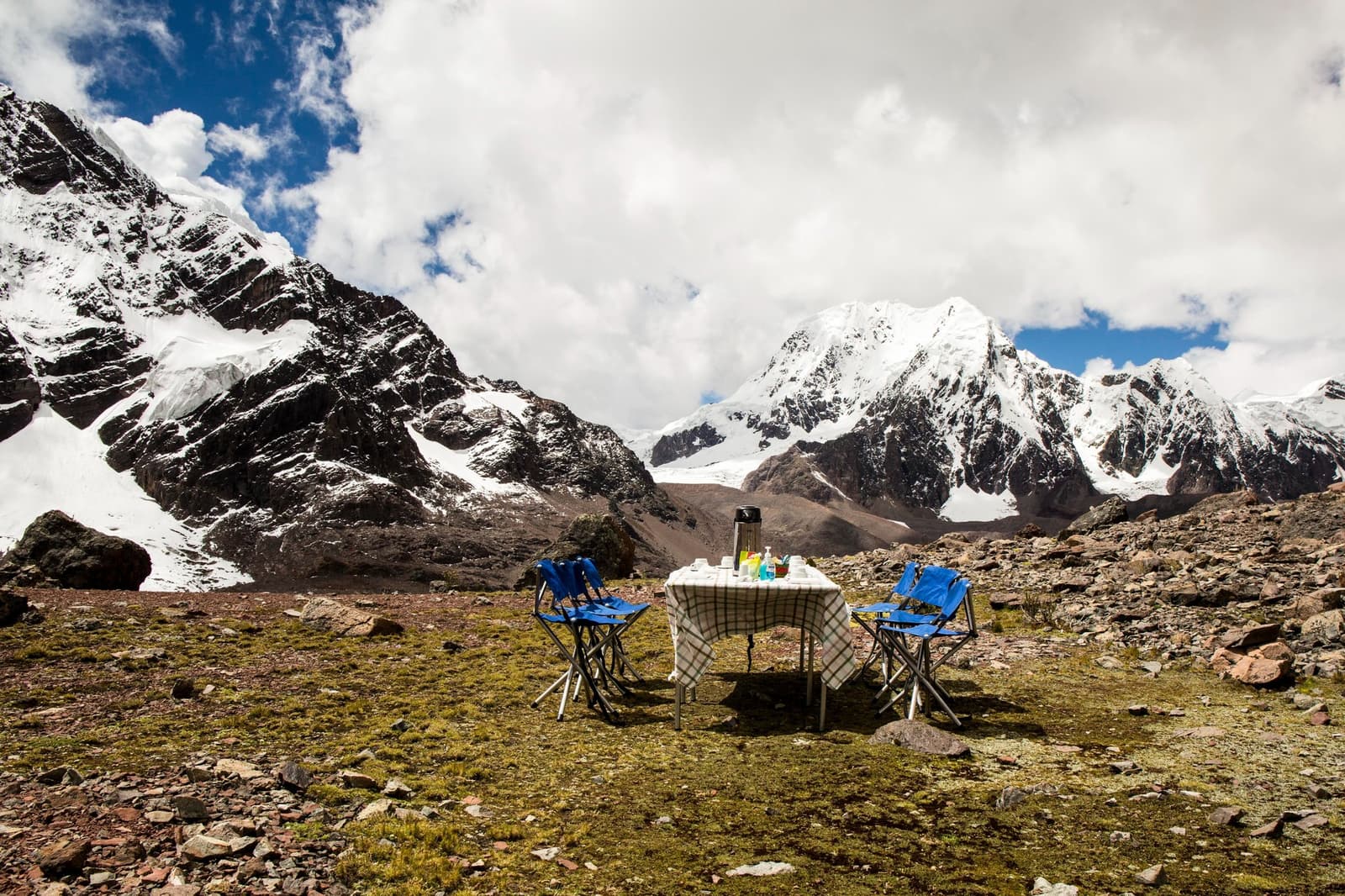 Lunch above 4,000 m, because the mountains can wait a few minutes. A folding table set with a checkered cloth, thermos, and refreshments surrounded by four blue camp chairs on rocky alpine terrain during the ausangate trek, with snow-capped Andean peaks rising under a partly cloudy sky
