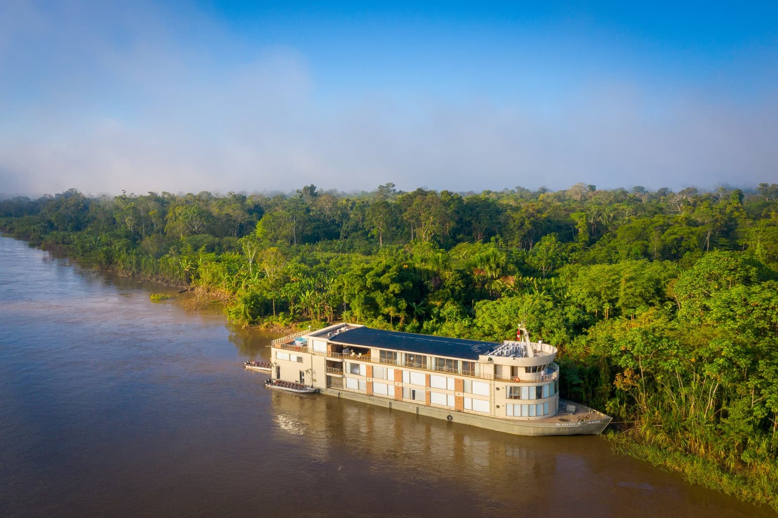 Quatro dias, um rio e uma floresta que parece não ter fim. Vista aérea do navio de cruzeiro Delfin Amazon atracado à margem de uma densa floresta tropical sob céu azul, com um pequeno bote partindo da popa nas águas pardas do rio