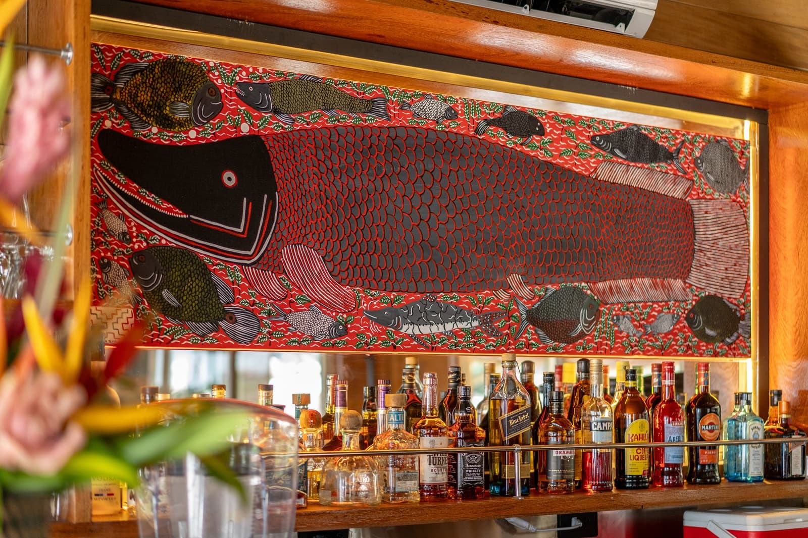 Arte do rio na parede, drinks à mão, o bar conta a história antes de você pedir o primeiro. Photo: Mario Gomi Painel de arte popular retroiluminado com um grande peixe escamoso rodeado por peixes menores em vermelho, preto e verde, exposto acima de um bar bem abastecido no cruzeiro Delfin na Amazônia, com garrafas de Campari, Kahlúa e Martini alinhadas na prateleira de madeira abaixo
