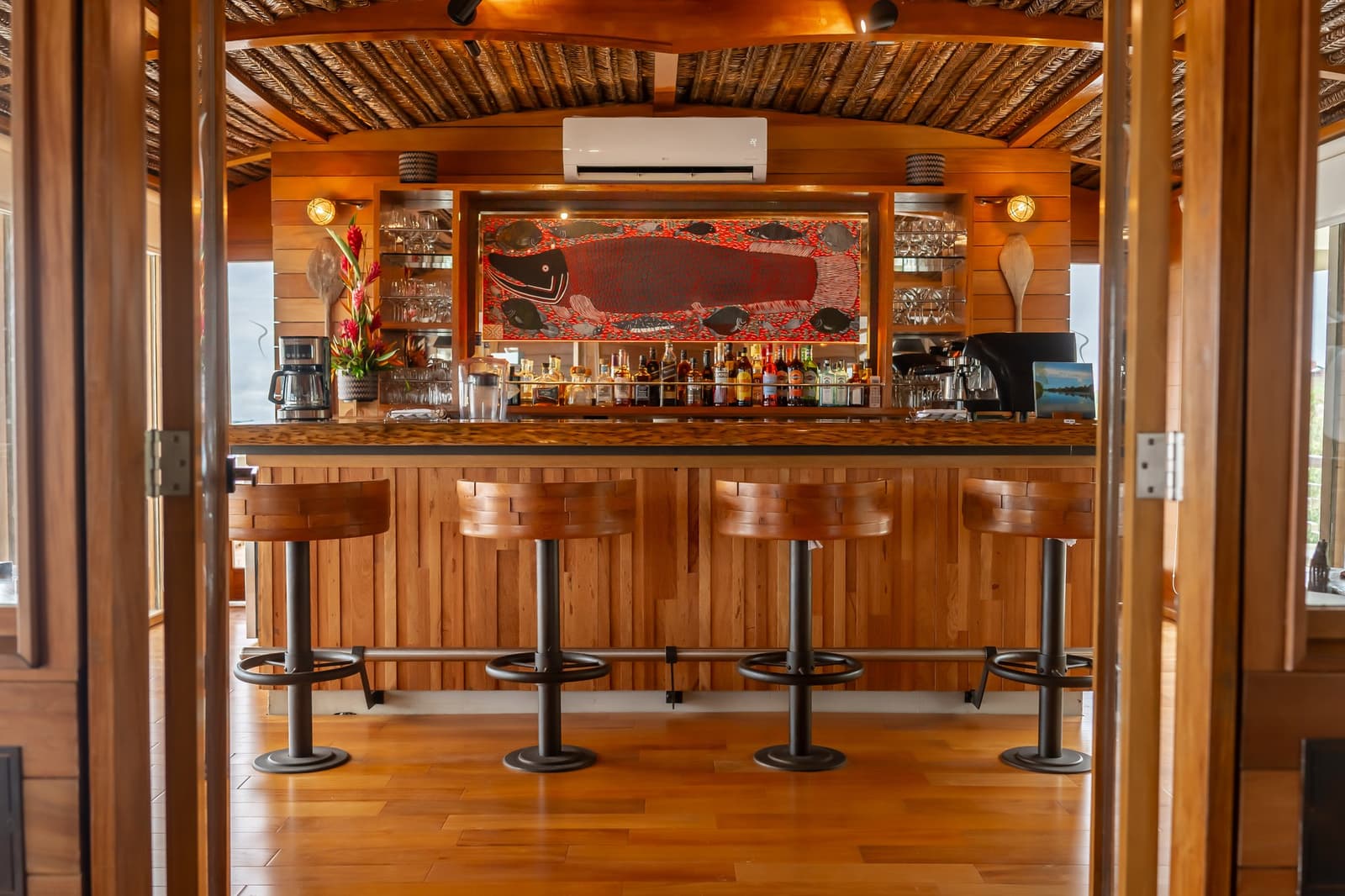 Um pisco sour antes do jantar, golfinhos-de-rio logo ali pela janela. Difícil reclamar. Photo: Mario Gomi Bar revestido de madeira do cruzeiro Delfin Amazon com quatro banquetas, prateleiras de bebidas bem abastecidas e uma obra de arte indígena com um peixe vermelho em destaque