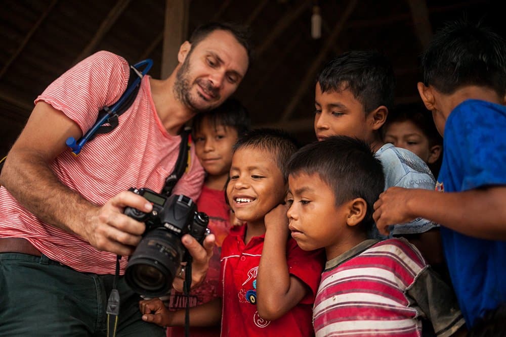 A foto na tela fez mais amizades do que qualquer guia turístico. Photo: : Rodrigo Rodrich Um viajante sorridente segurando uma câmera DSLR Nikon mostra a tela para um grupo de crianças animadas durante uma visita comunitária no cruzeiro Delfin pelo Amazonas no Peru