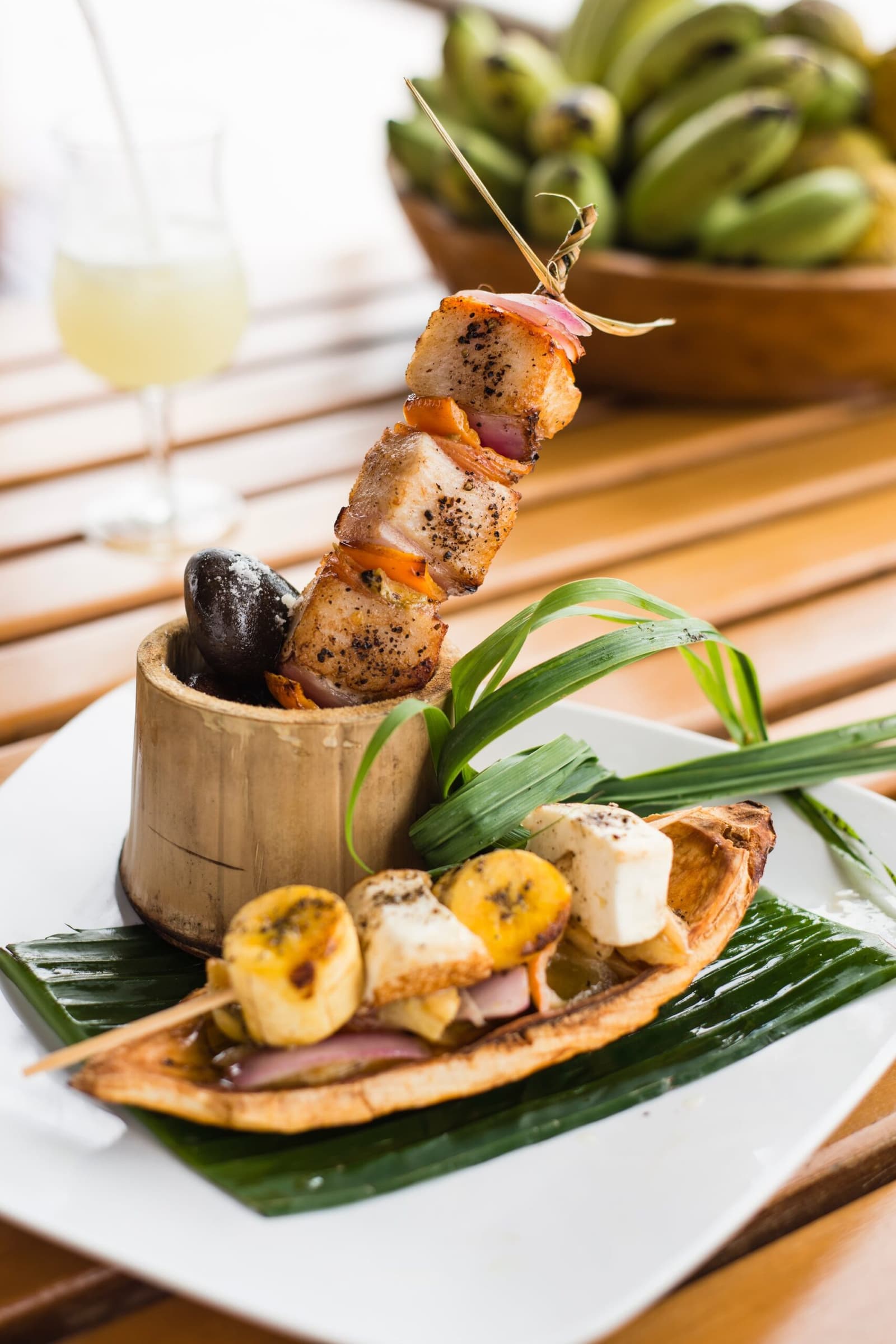 Ingredientes locais, um copo de bambu e uma cozinha que leva os dois a sério. Photo: MARIO GOMI Espeto de carne grelhada e legumes apoiado verticalmente em um copo de bambu, servido sobre folha de bananeira ao lado de um espeto de banana-da-terra caramelizada e queijo, como parte da experiência gastronômica do cruzeiro Delfin na Amazônia