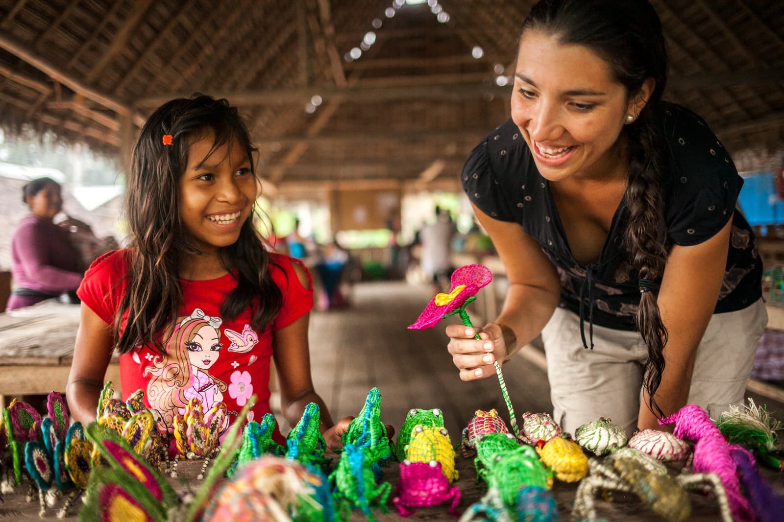 Os sapos são de palha trançada; os sorrisos, absolutamente genuínos. Photo: er: Rodrigo Rodrich Uma menina sorridente e uma visitante observam artesanatos coloridos trançados — sapos, flores e pequenos animais — dispostos sobre uma mesa no mercado comunitário de um vilarejo ribeirinho, num pavilhão com teto de palha, durante uma parada do cruzeiro Delfin Amazon.