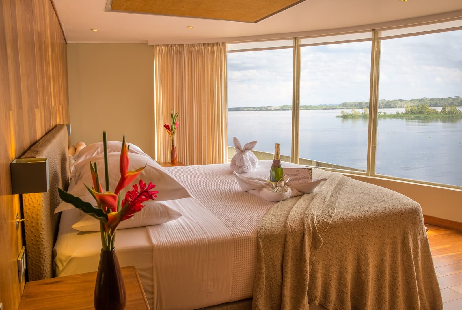 Champanhe na cama e o Amazonas pela janela. A viagem começa bem. Photo: DIEGO PEREZ Uma cabine bem decorada no cruzeiro Delfin Amazon com cama de casal, escultura de toalha e garrafa de champanhe, flores tropicais na mesinha de cabeceira e janelas do piso ao teto com vista para um rio largo e ilhas cobertas de vegetação.