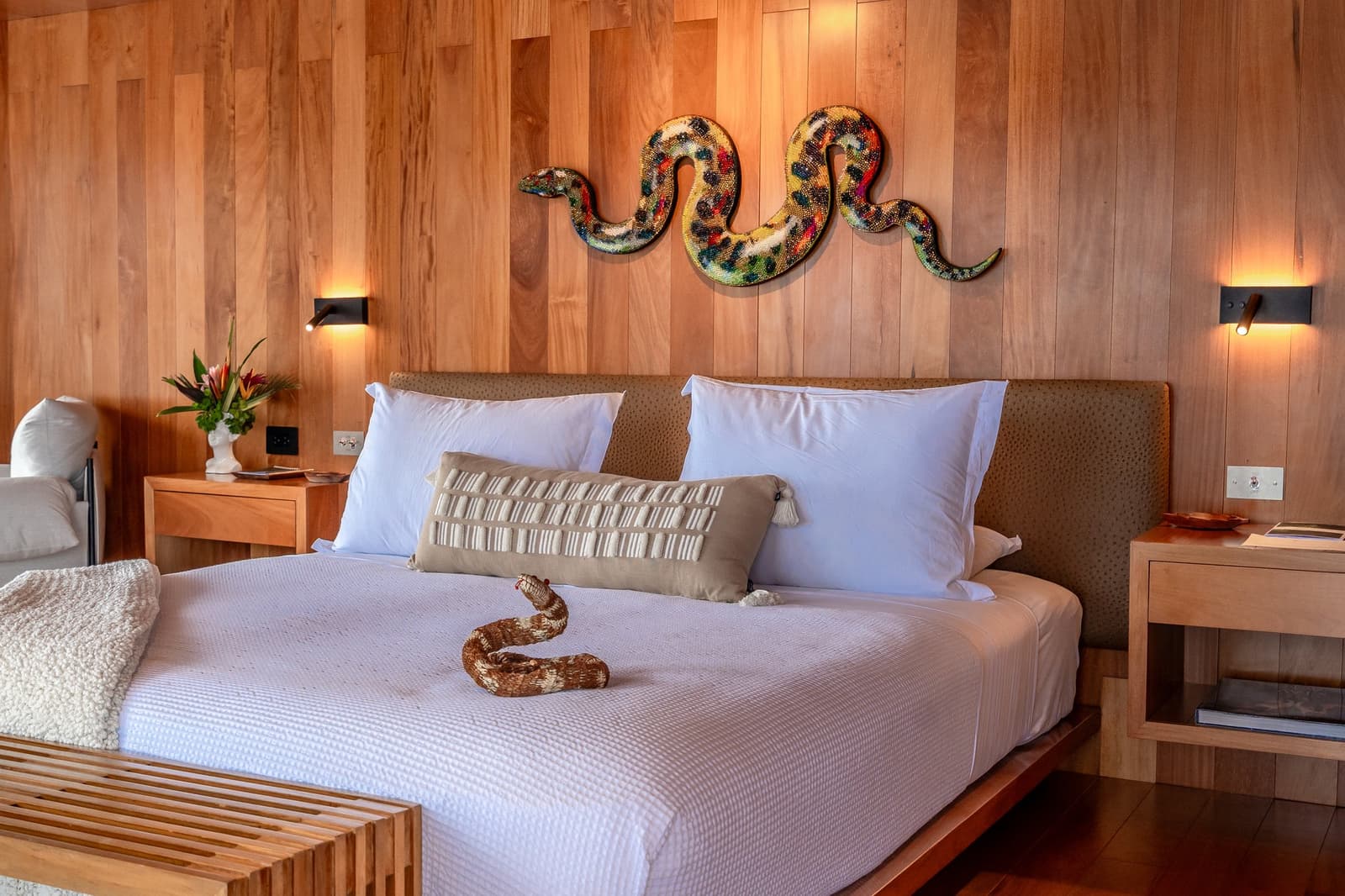 Duas cobras, zero reclamações, a suíte tem personalidade própria. Photo: Mario Gomi Cama king-size com roupa de cama branca em uma suíte de painéis de madeira quente no cruzeiro Delfin Amazon, com uma escultura mural de serpente em mosaico colorido acima da cabeceira e uma figura de serpente trançada sobre a cama