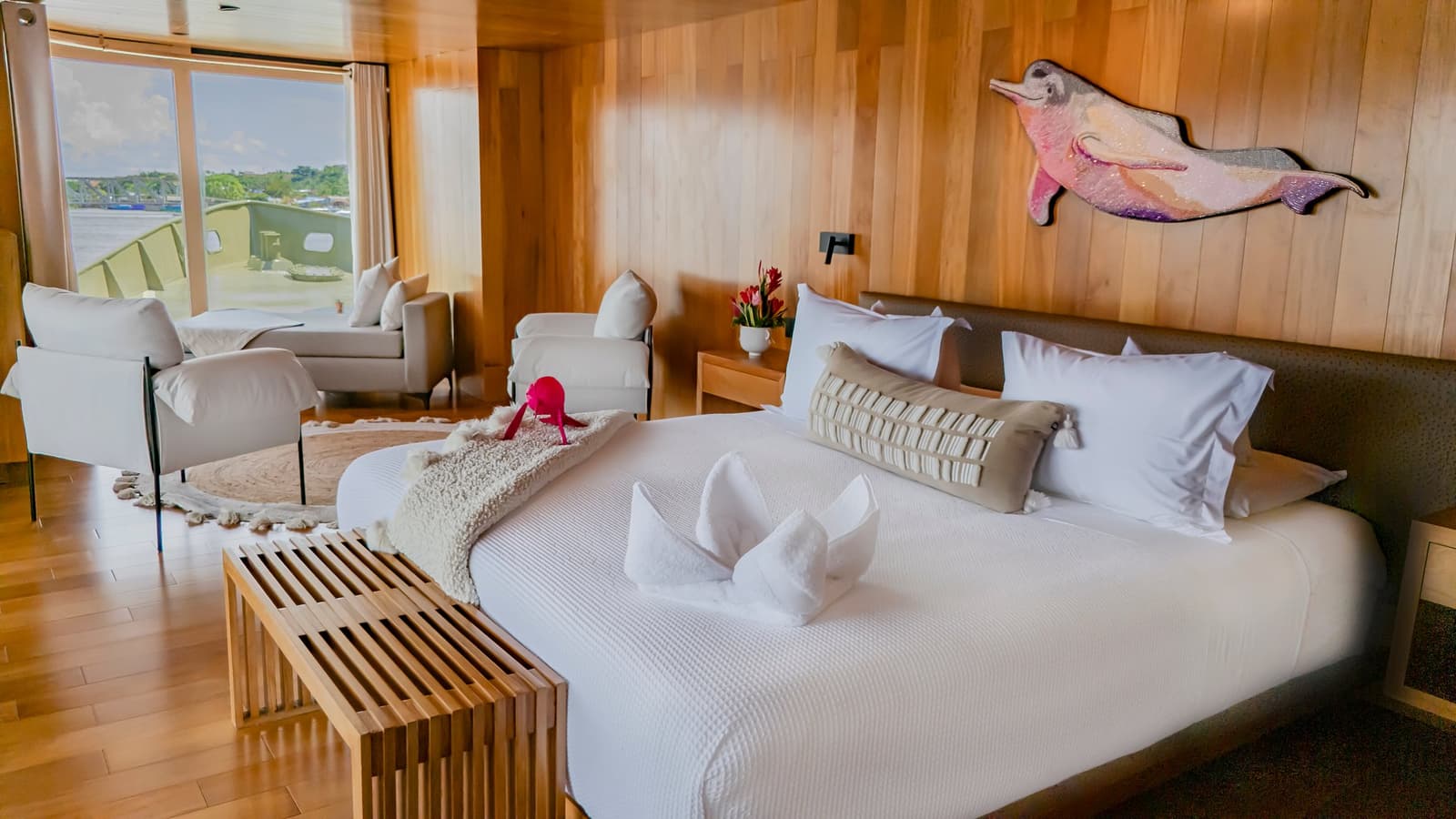A arte na parede já avisa: você está no coração da Amazônia. Cabine espaçosa no Delfin Amazon Cruise com cama king em roupa de cama branca, origami de toalhas dobradas, escultura decorativa de boto-cor-de-rosa na parede, painéis de madeira aquecidos e vista para o rio pelas grandes janelas