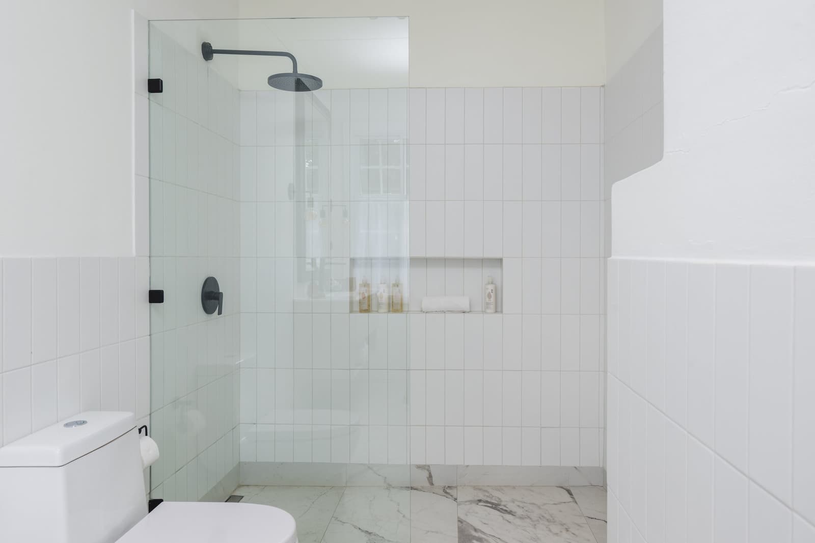 A proper rinse after a long day on the steppe, no complaints here. Photo: CARLOS DIAZ White-tiled walk-in shower with a frameless glass panel, matte black rainfall showerhead and fixtures, built-in niche stocked with toiletries, and marble-effect floor tiles in a private bathroom at Estancia Cerro Guido