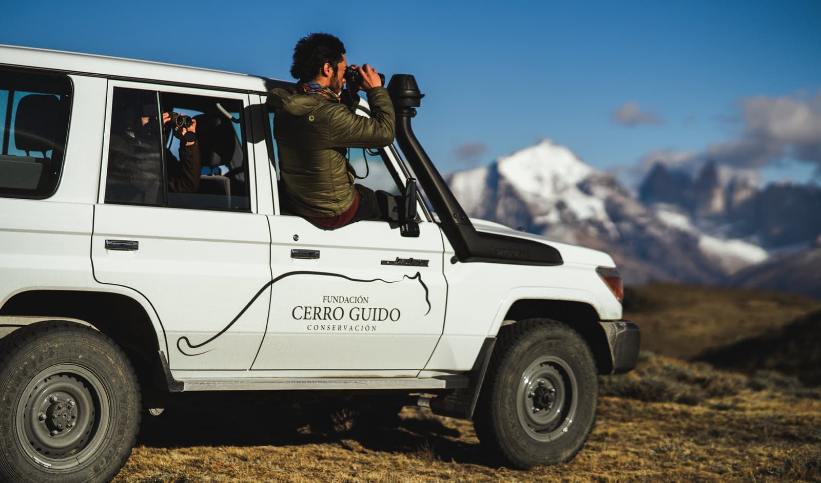 Binoculars out, engine off. The steppe does the rest. A guide leans out of a Fundación Cerro Guido conservation 4x4 vehicle scanning the steppe with binoculars, snow-capped peaks visible in the background under a clear blue sky