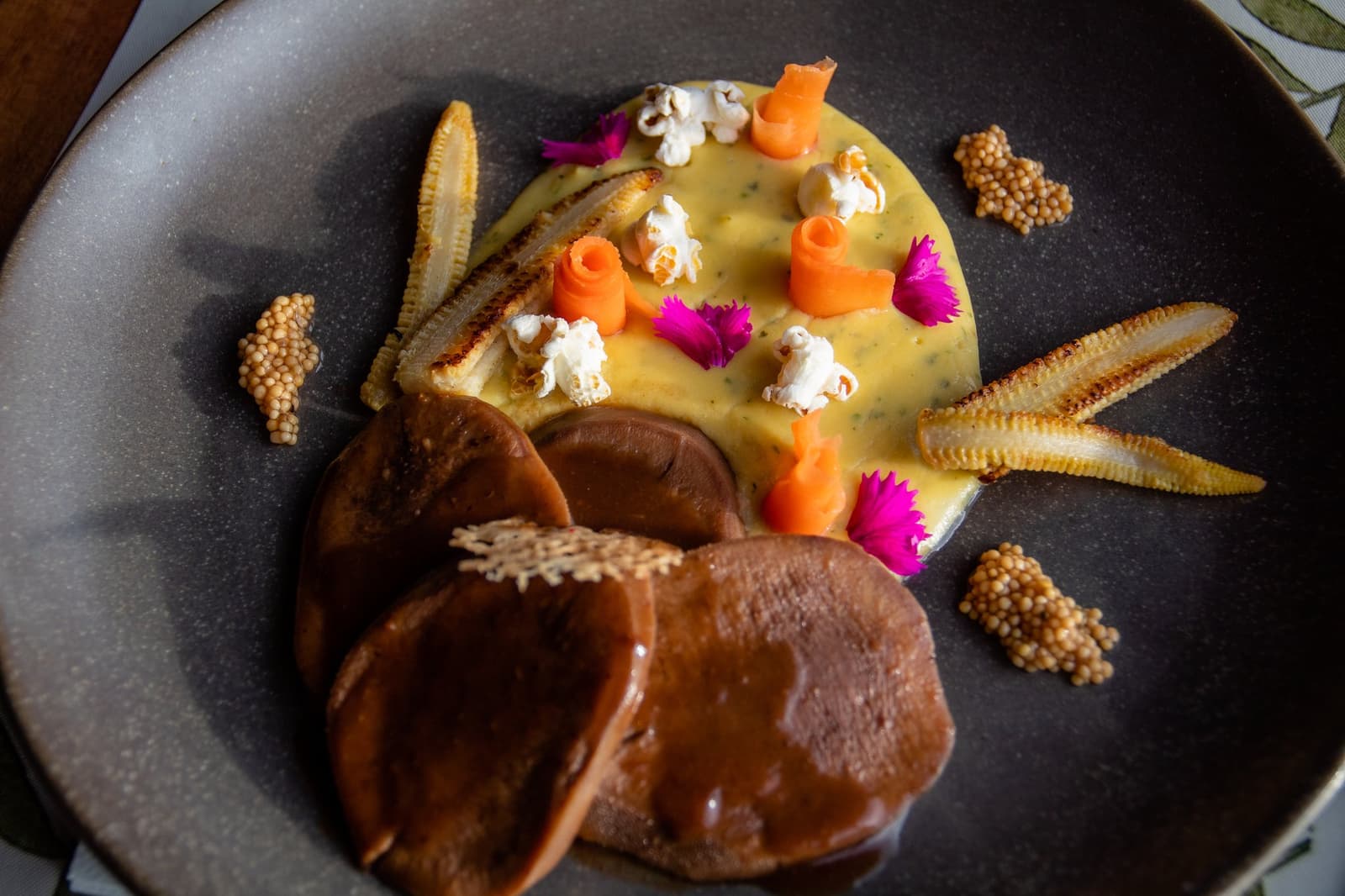 Ranch cooking with a creative edge, well past the lamb-and-potatoes brief. An artfully plated dish at Estancia Cerro Guido featuring glazed rounds in a golden herb sauce, garnished with popcorn, carved carrot curls, pink edible flowers, crispy baby corn, and puffed grain clusters on a dark ceramic plate