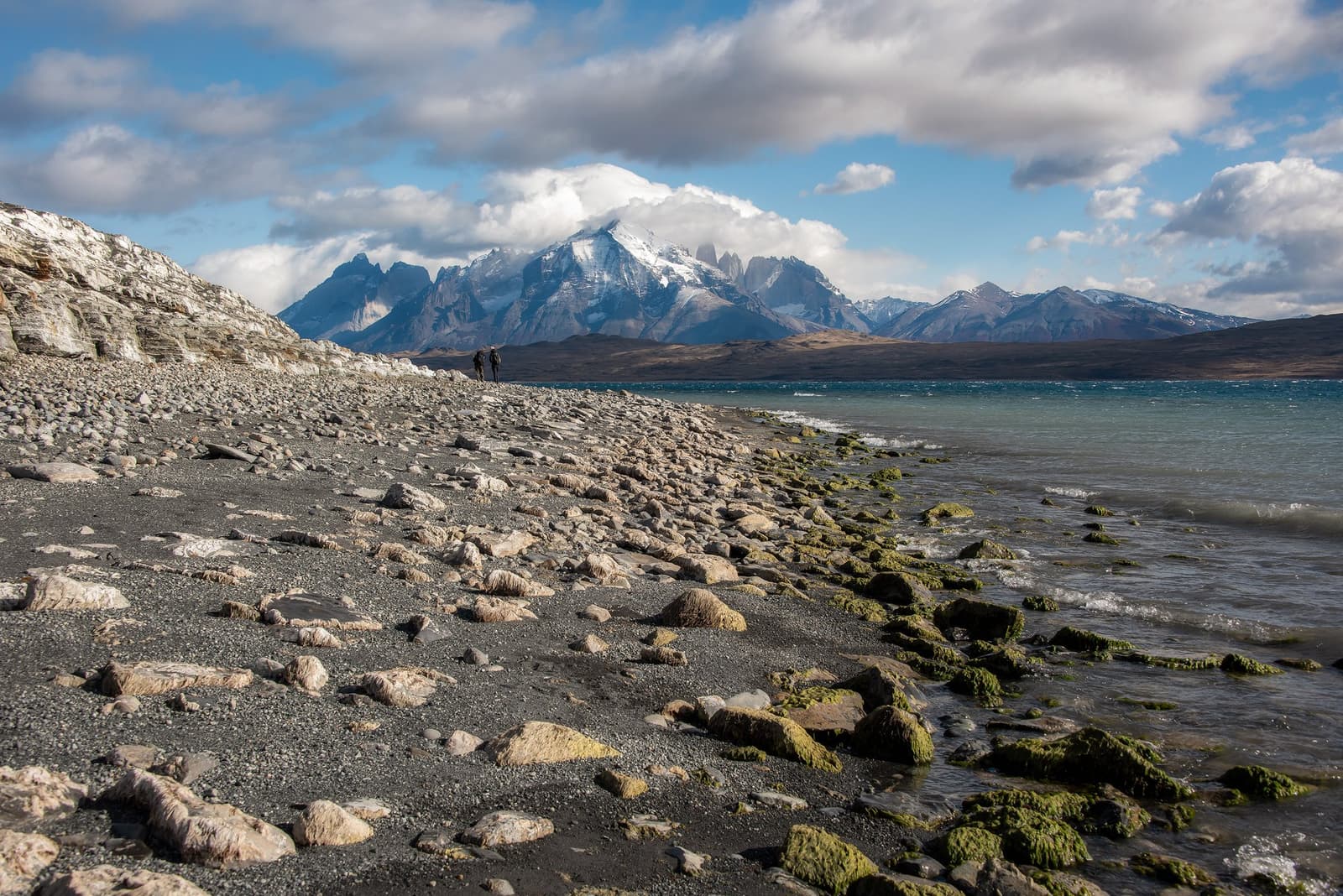The lake keeps the mountains honest, and the scale keeps you humble. Two hikers walk along a rocky, moss-edged lakeshore at estancia cerro guido, with the snow-dusted granite towers of the Paine Massif rising under a cloudy sky in the distance