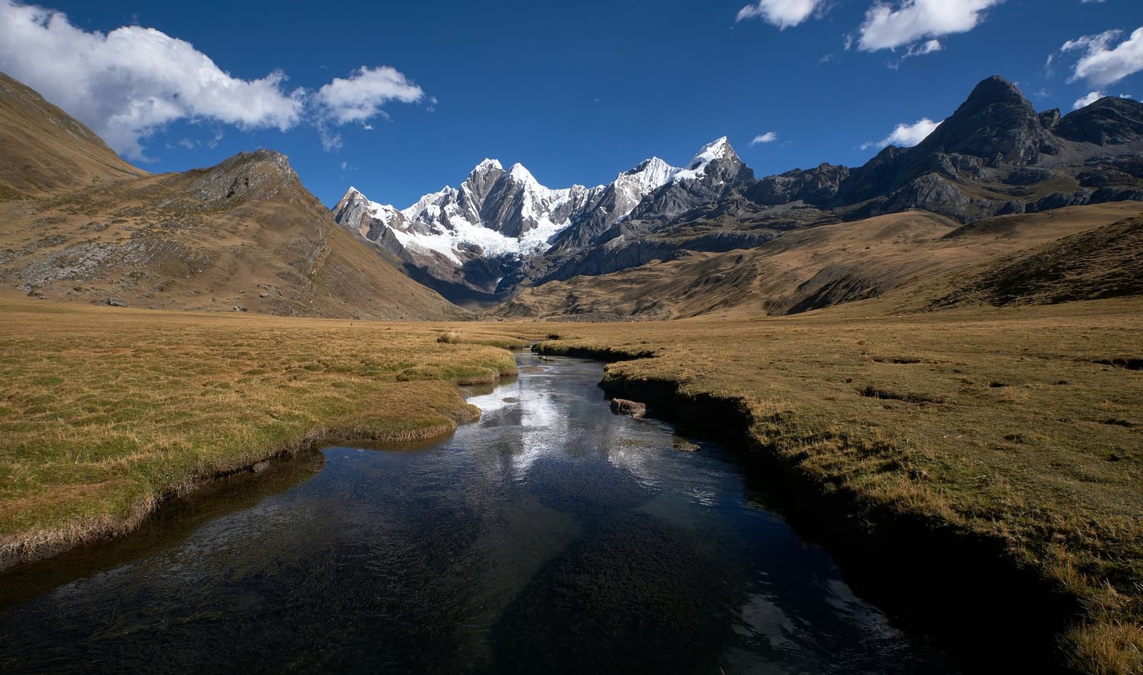 Cold water, open sky, and a valley that just keeps going. A clear stream winds across a wide high-altitude puna grassland on the huayhuash circuit, reflecting the deep blue sky as it leads the eye toward a wall of snow-capped Andean peaks rising at the head of the valley