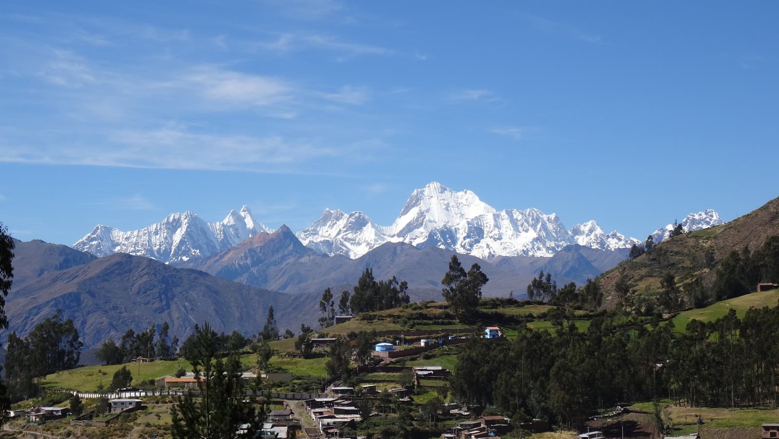 The last town before the mountains take over completely. A small Andean village with terracotta-roofed houses and eucalyptus trees sits in a green valley, with the snow-capped peaks of the cordillera huayhuash filling the horizon under a clear blue sky.
