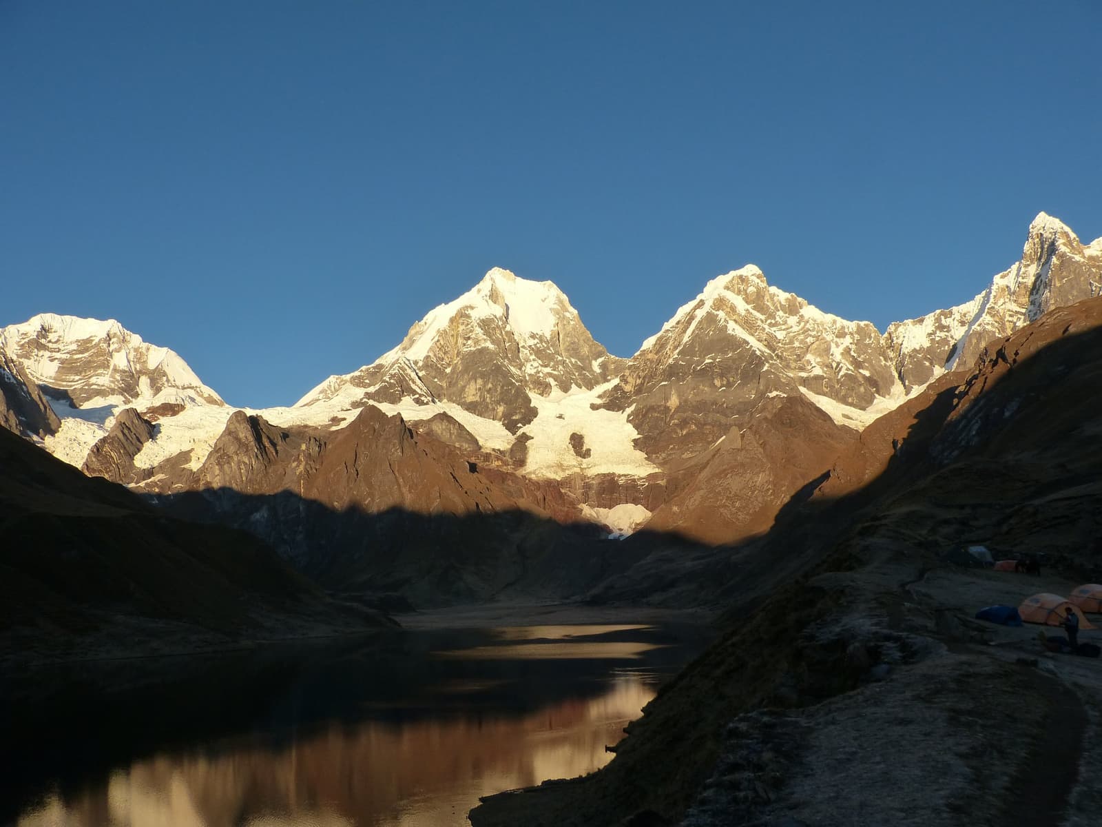Camp on, peaks on, coffee hopefully on. Golden early light on snow-capped peaks of the huayhuash circuit reflected in a still glacial lake, with colorful expedition tents pitched on the rocky shore in the foreground