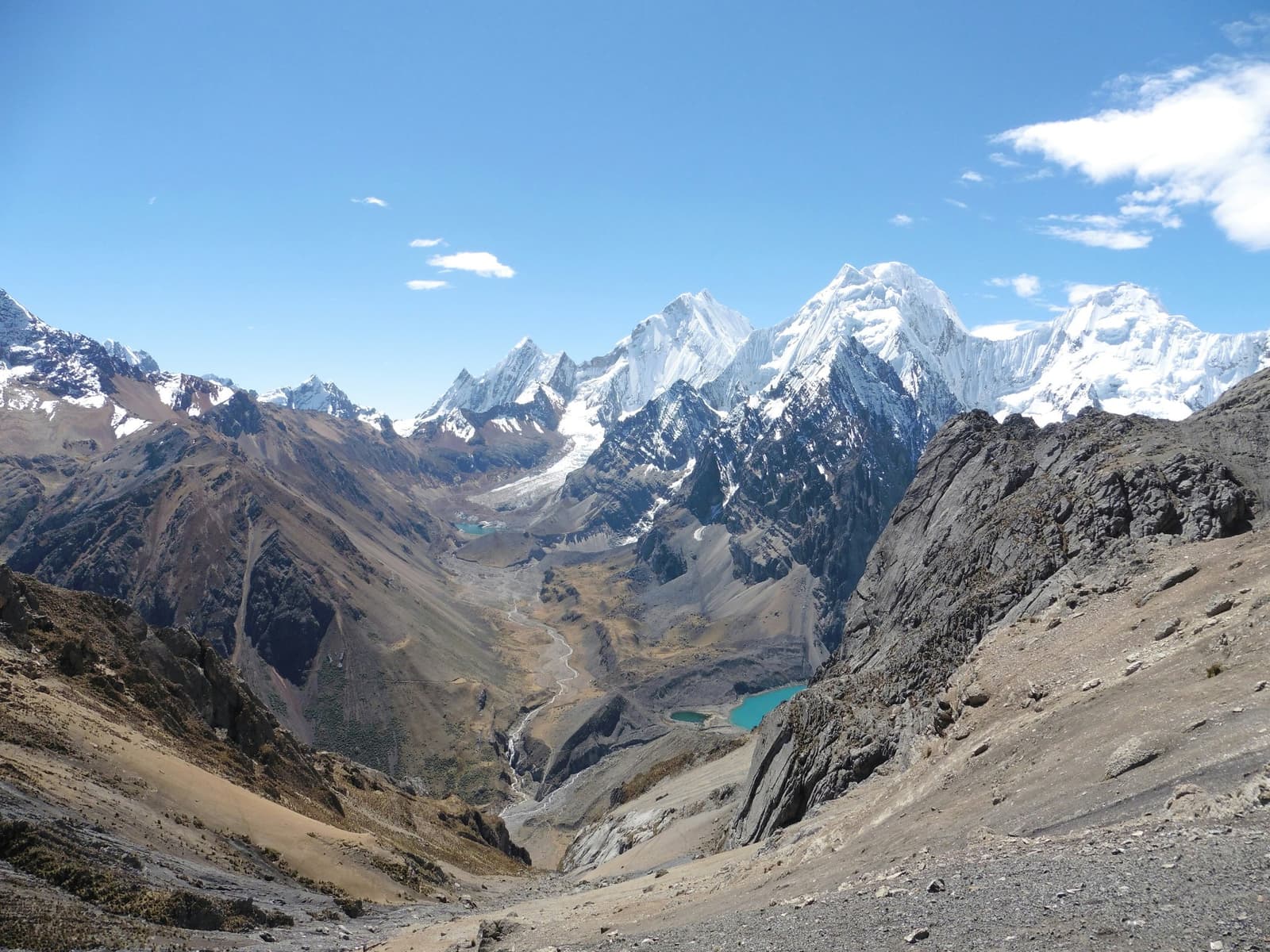 Two lakes, one valley, and a glacier that's been busy for centuries. Wide aerial view of the huayhuash circuit looking down a high-altitude glacial valley with two turquoise lakes, a braided meltwater stream, and snow-capped Andean peaks rising against a clear blue sky