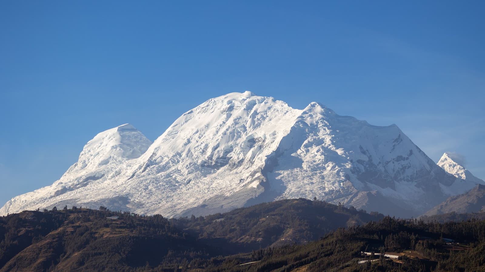 Serra enorme, céu aberto, e a altitude avisando que está lá. Photo: MAREK PIWNICKI Um amplo maciço coberto de neve com vários cumes glaciados se ergue acima de colinas arborizadas sob um céu azul limpo, fotografado durante uma caminhada de aclimatização perto de Huaraz, no Peru.