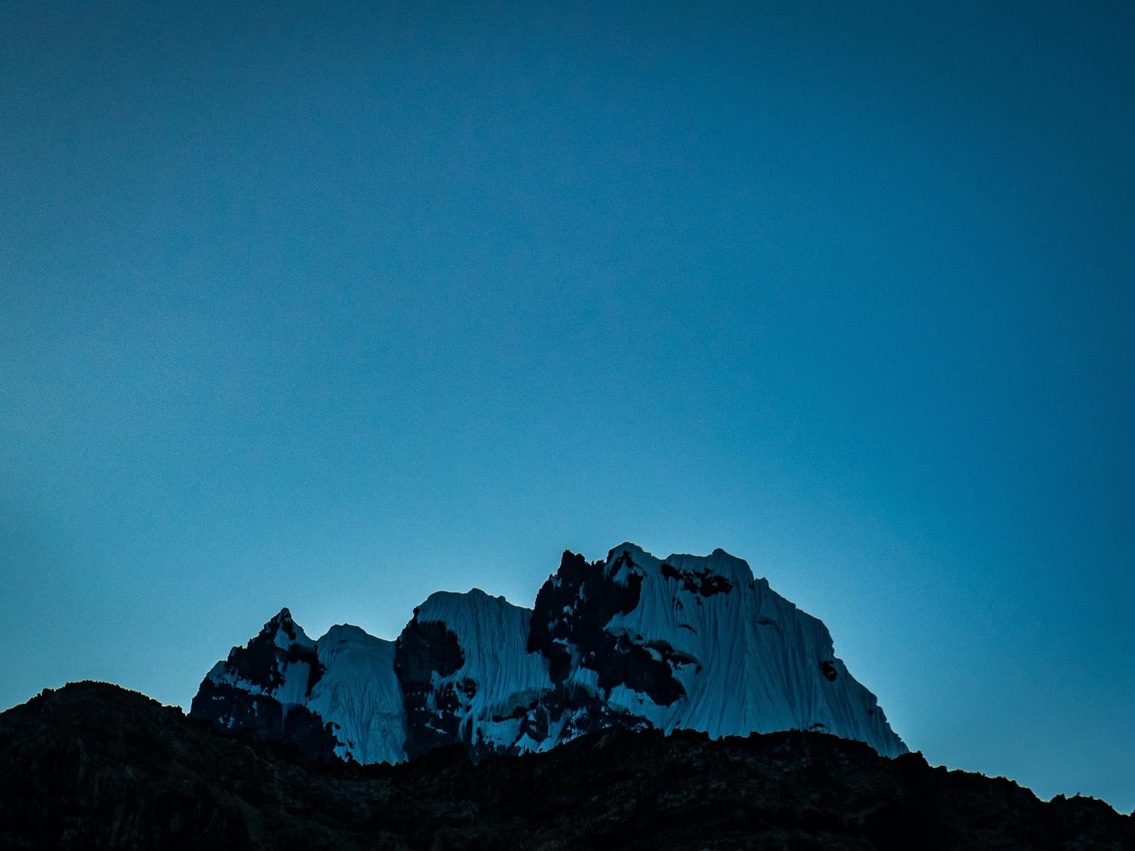 That cold blue hour before dawn, and the glaciers are already awake. Snow-draped rocky summit of the huayhuash circuit glowing faintly against a deep blue twilight sky, with dark forested ridgelines silhouetted in the foreground