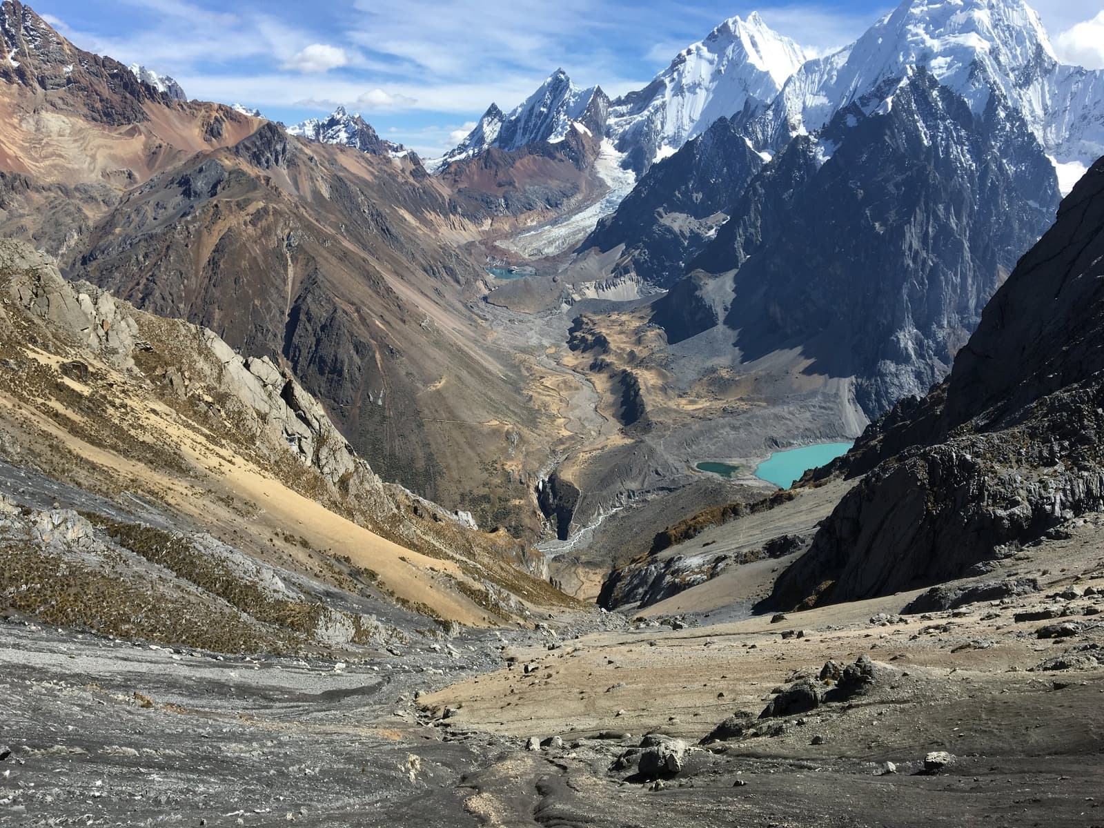 That electric blue lake earned its color the hard way, one glacier at a time. Wide glacial valley on the huayhuash circuit viewed from high altitude, with a turquoise glacial lake gleaming on the right, a braided meltwater stream winding down the center, and snow-capped peaks rising sharply under a blue sky