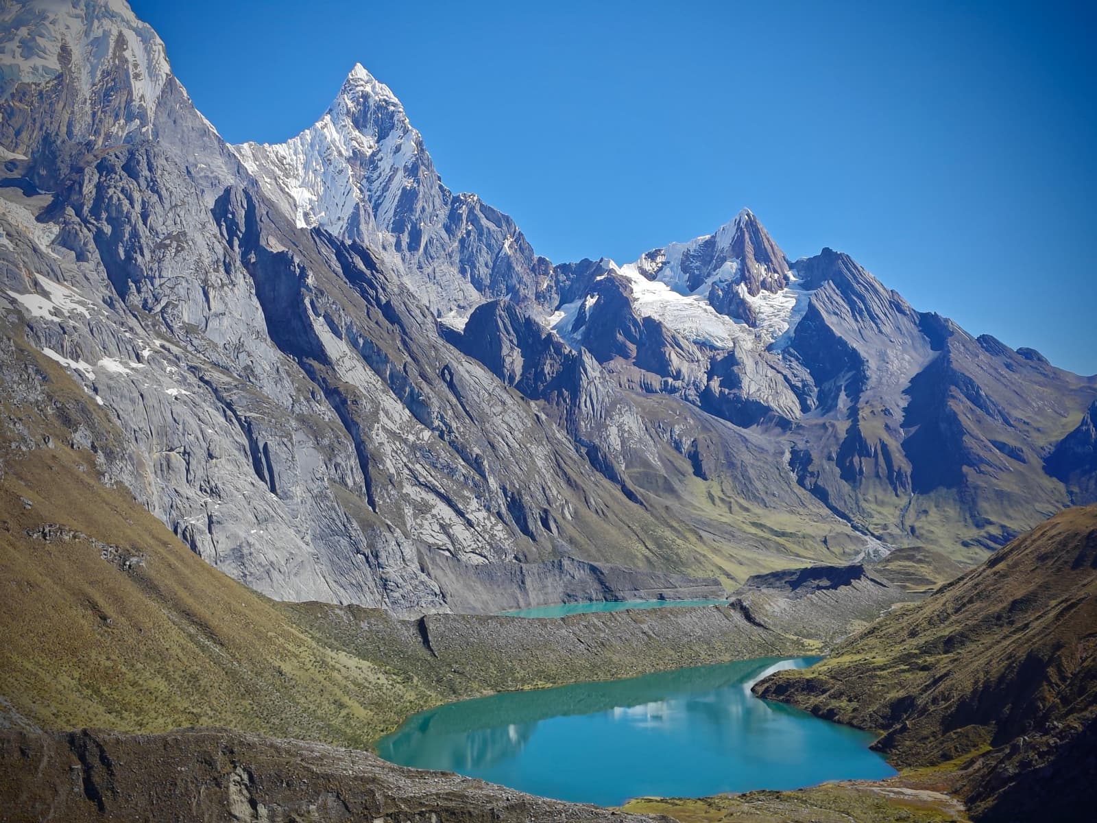 Three lakes, one after another, like the mountains couldn't stop showing off. Three turquoise glacial lakes sit in a high Andean valley on the huayhuash circuit trek, ringed by jagged snow-capped granite peaks and steep rocky cliffs under a deep blue sky