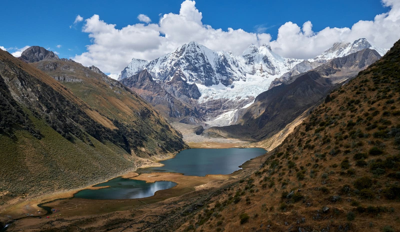 Two lakes, one glacier, and the whole valley laid out like it's showing off. Aerial view of two turquoise glacial lakes in a high-altitude Andean valley on the huayhuash circuit, with snow-capped peaks and a hanging glacier rising behind them under a blue sky with white clouds