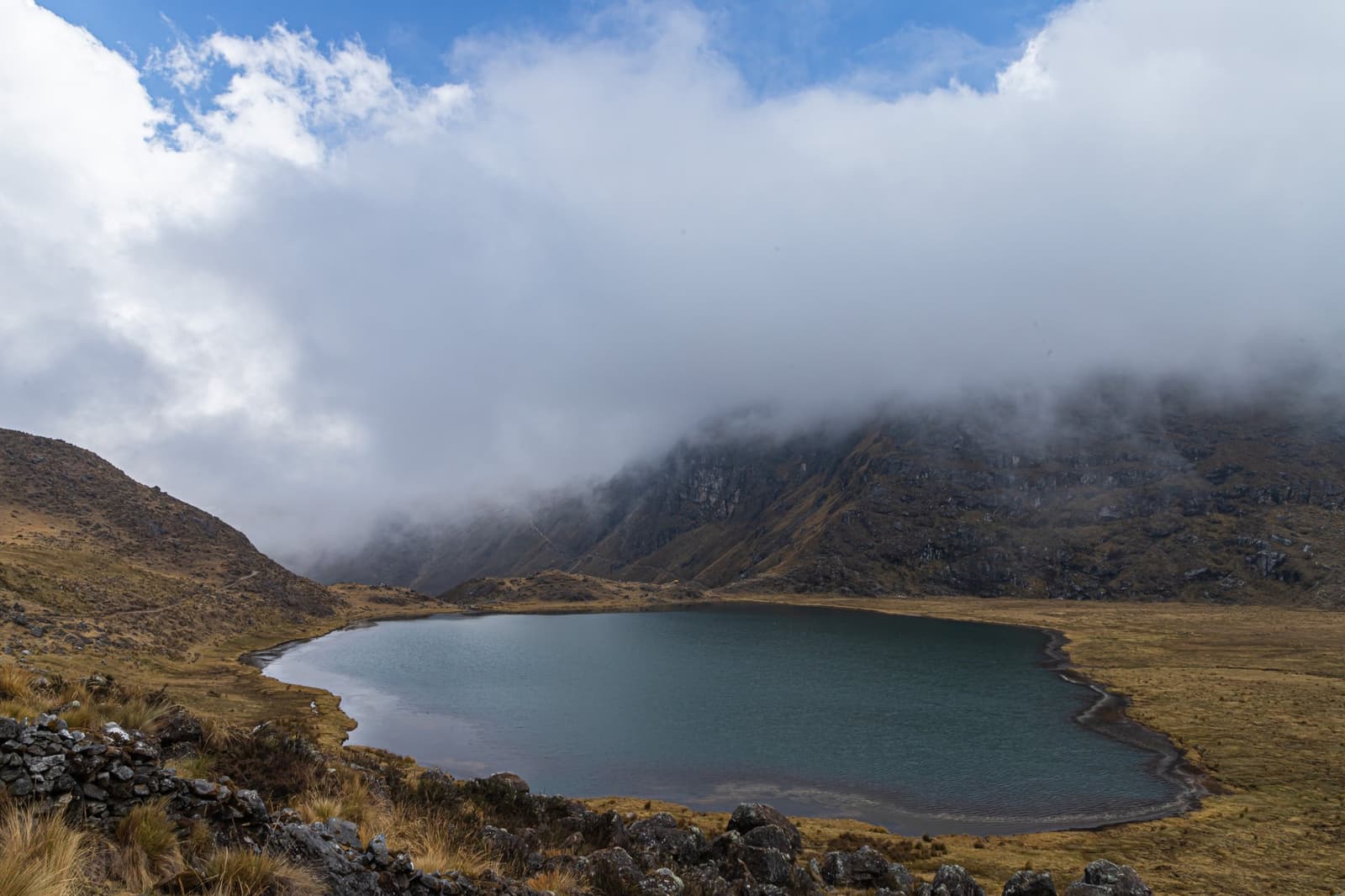 Clouds doing exactly what they want, right on schedule. Photo: GUILLERMO MIGUEL CARLOS GOMEZ A round high-altitude lake with teal water sits in an open puna valley on the huayhuash trek, with low clouds rolling over the rocky ridge behind it and golden ichu grass covering the hillsides
