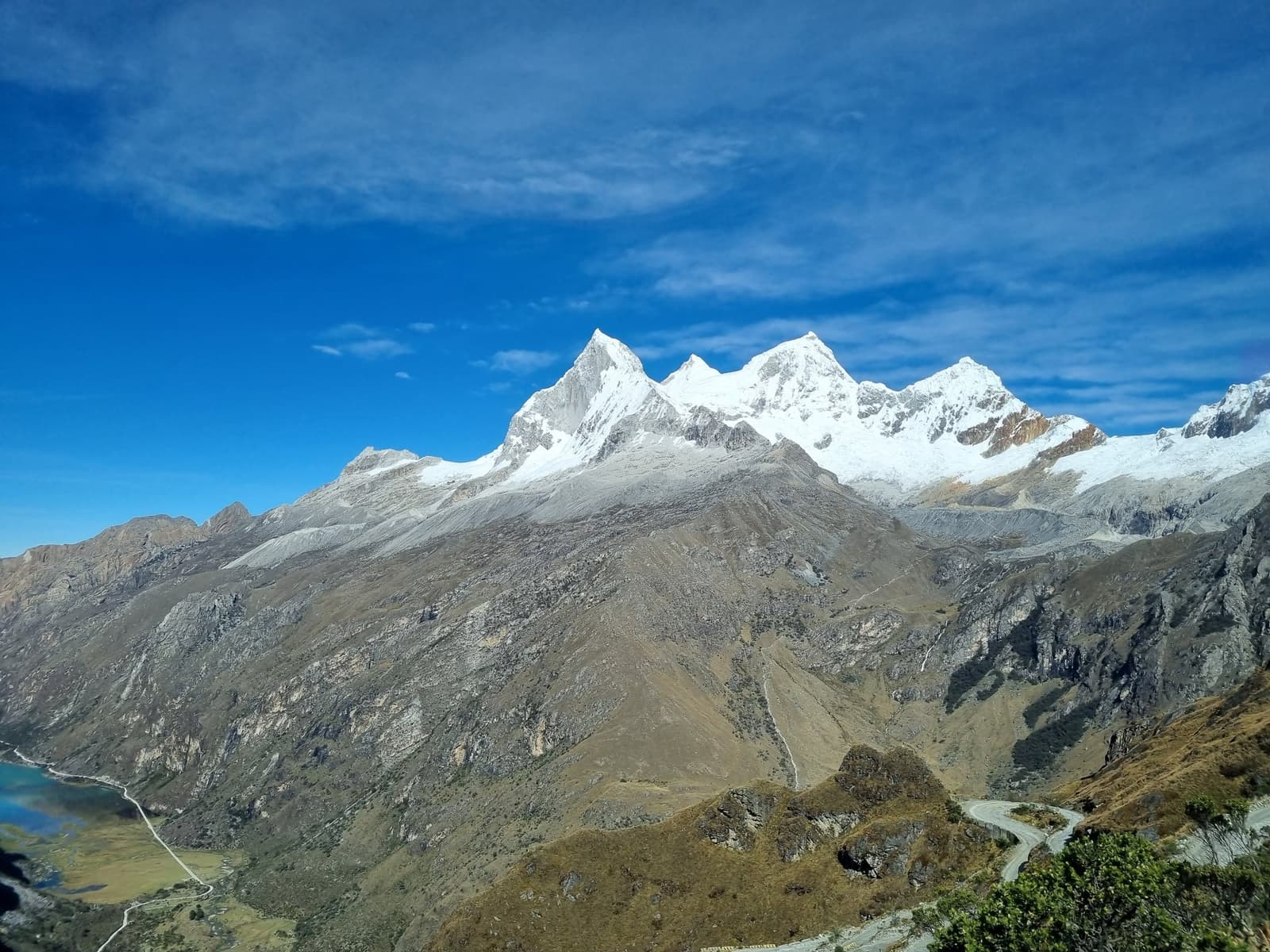 Montanhas enormes, uma estrada fina, silêncio total. É assim que começa a aventura. Picos cobertos de neve da Cordilheira Huayhuash se erguem acima de um vale de alta altitude acidentado, com uma estrada sinuosa e um lago glacial visível ao longe sob um céu azul intenso