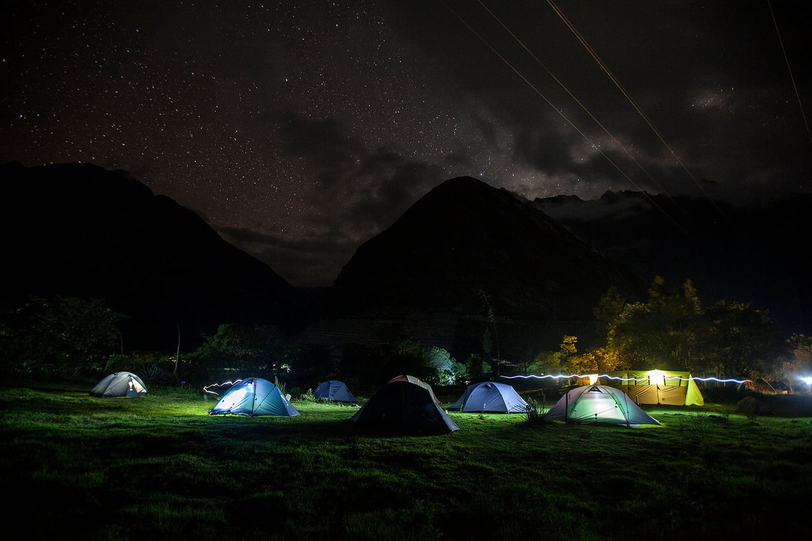 Your tent is warm, the stars are out, and tomorrow the real climbing begins. A cluster of illuminated tents pitched on a grassy flat during an Inca Trail to Machu Picchu trek, with dark mountain silhouettes and a star-filled sky overhead