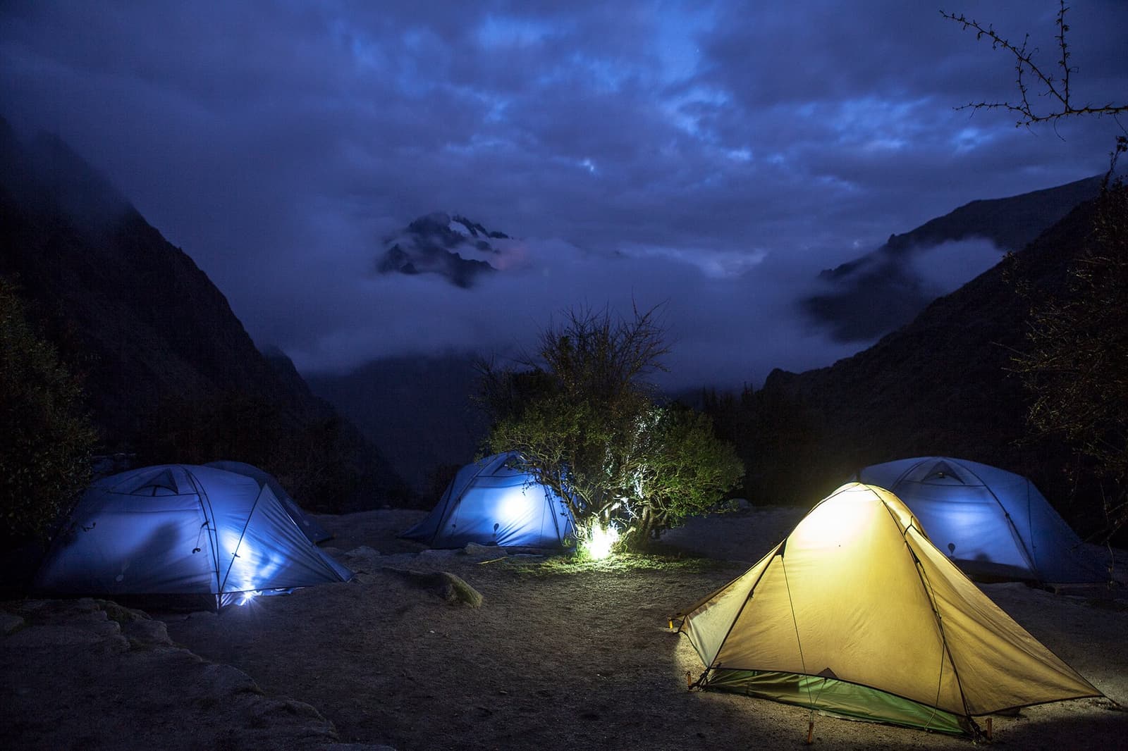 Sleep here, and the mountains are your only neighbors. Three illuminated tents glowing in blue and gold at a high-altitude campsite on the Inca Trail to Machu Picchu, with misty Andean mountain peaks and a dramatic cloudy night sky above