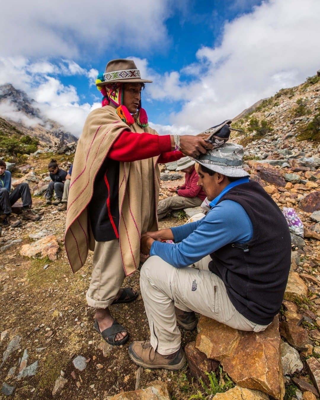 A small gesture, a hat exchanged, and suddenly you're part of something much older than tourism. A man in traditional Andean dress with colorful tassels and a woven poncho places a textile hat on a seated trekker during a machu picchu adventure tour, with a rocky mountain landscape and blue sky behind them.
