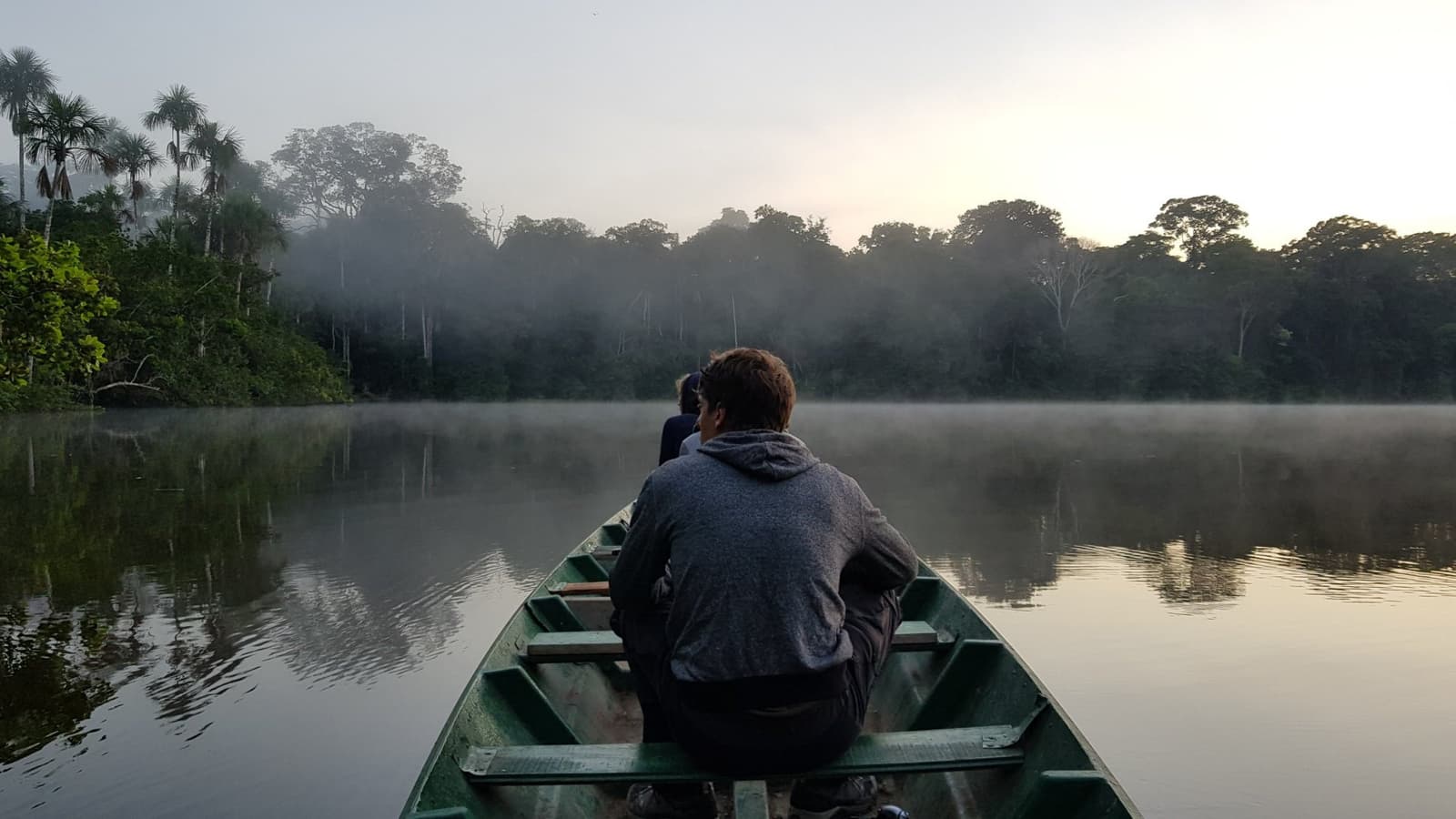 Five AM, mist on the water, and nobody is complaining about the early alarm. Photo: Pereira Two travellers sit at the bow of a green canoe gliding across a glassy oxbow lake on a Machu Picchu and Amazon tour, with morning mist rising off the water and dense rainforest reflected on the surface