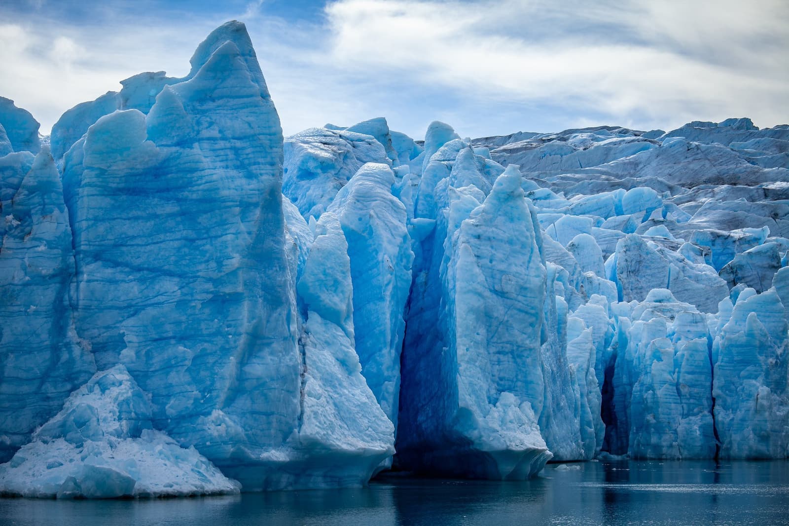 Ancient ice, compressed over centuries into a blue that no screen does justice. Towering blue ice seracs at a glacier terminus rising directly from dark water on the self-guided O Circuit in Torres del Paine, with deep crevasses splitting the ice wall under a cloudy sky
