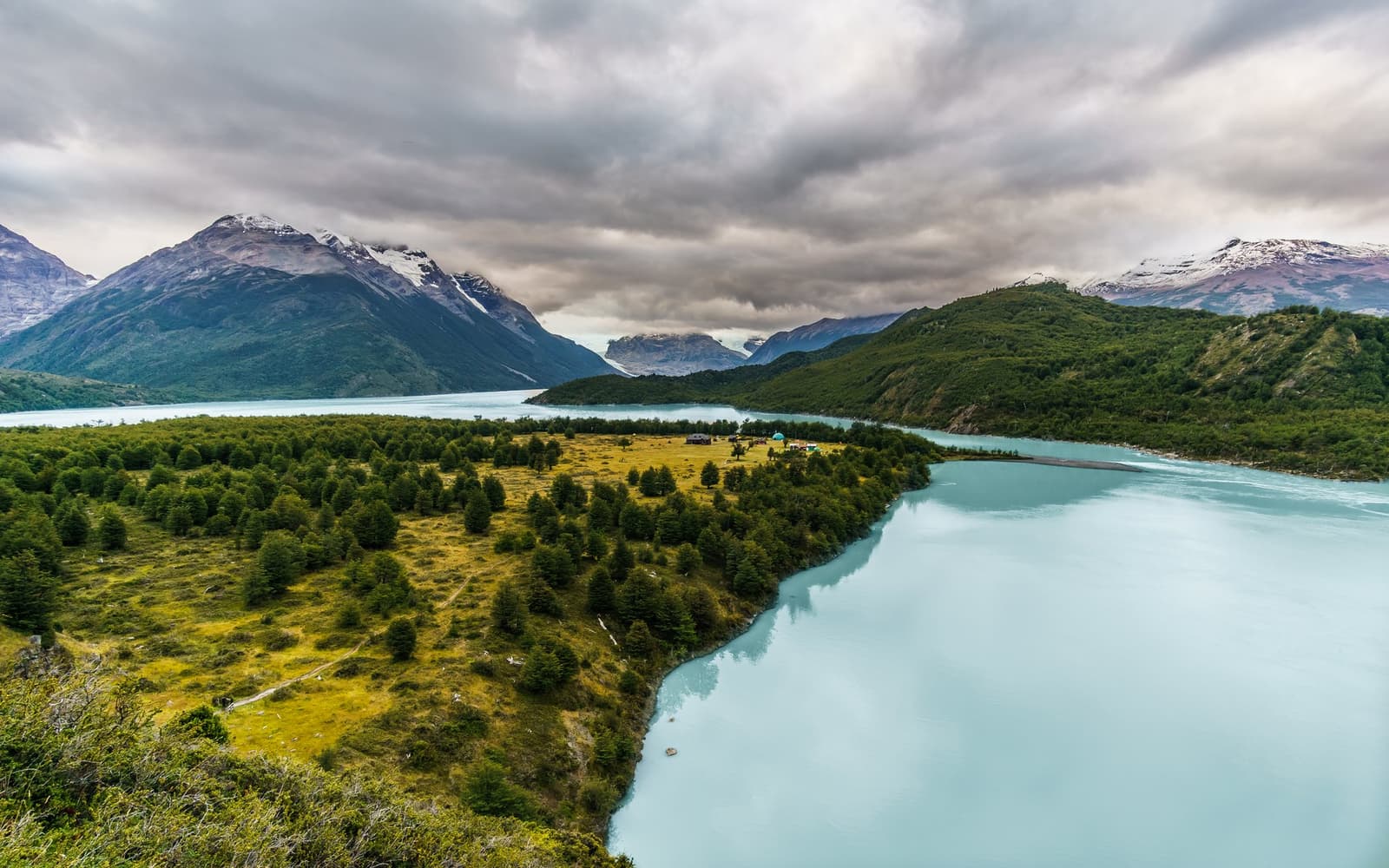 That campsite tucked into the trees is yours for the night. Worth every step. Aerial view of a glacial turquoise lake curving around a forested peninsula on the self-guided O Circuit, with snow-capped mountains and overcast skies in the background and a small campsite visible among the trees