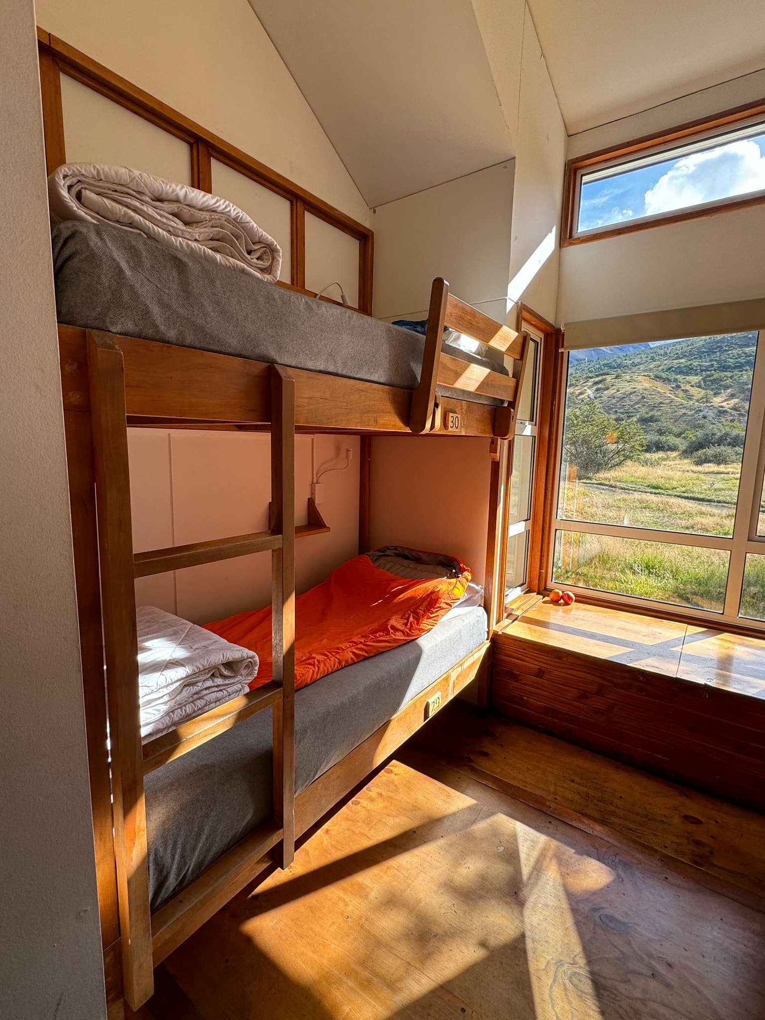 Afternoon sun on the lower bunk, hills waiting just outside the glass. Wooden bunk beds with orange sleeping bags in a self-guided O Circuit refugio room, large windows overlooking green Patagonian hillsides and open grassland