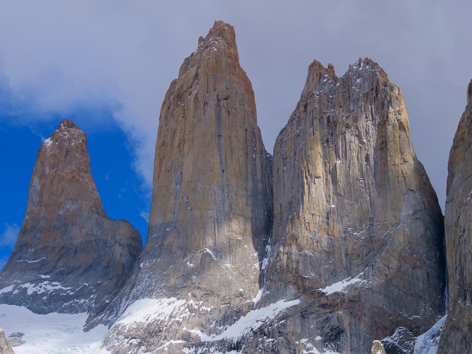 Three billion years of geology, no filter needed. Three sheer granite towers of Torres del Paine rise steeply above a snow-dusted base on the self-guided O Circuit, with warm ochre and grey rock faces filling the frame under a partly cloudy sky