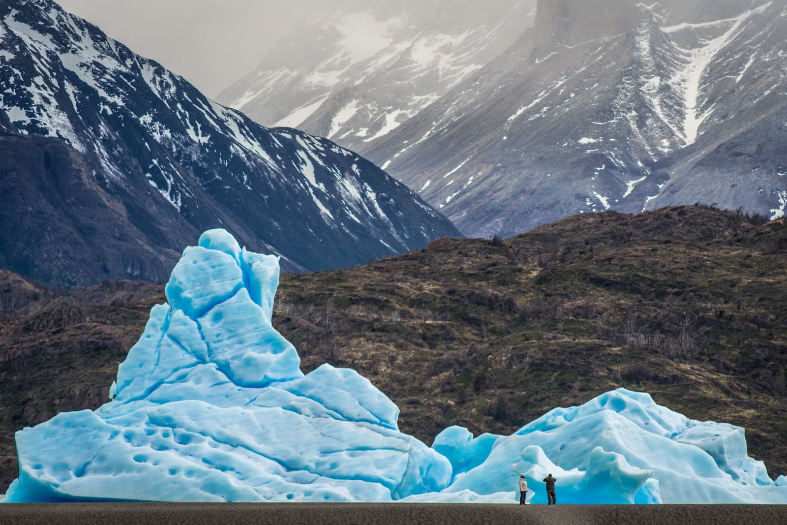 Two people, one iceberg, no sense of scale until you zoom in. Two travellers standing at the base of a massive blue iceberg on the o trek patagonia route, with snow-streaked Andean peaks rising through cloud in the background