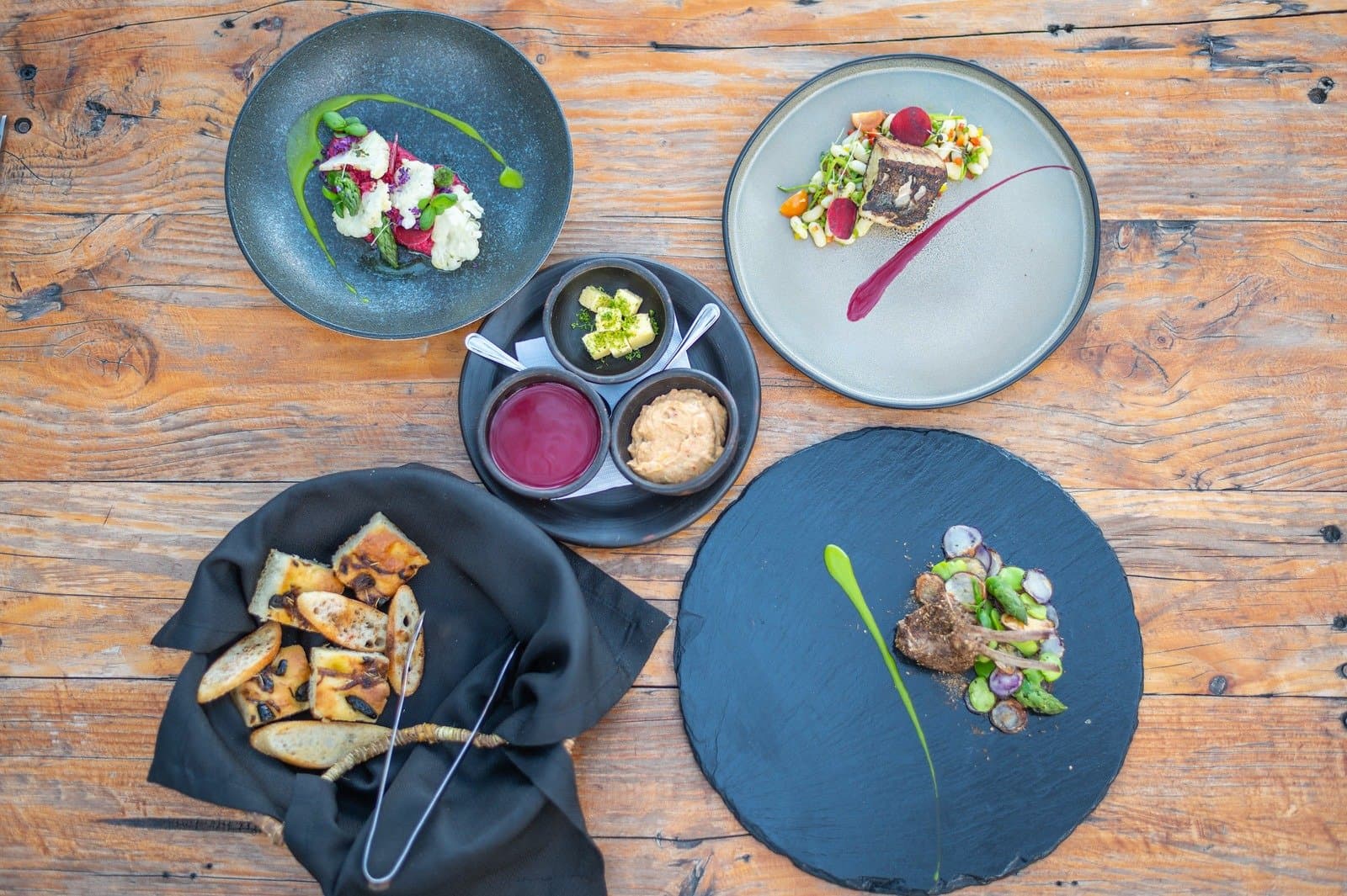 After 12 hours on the pass, this table earns every bite. Overhead flat-lay of five plated dishes and a bread basket on a rustic wood table at EcoCamp Patagonia, showcasing creative small-portion cuisine with colourful sauces, grilled bread, and fresh vegetable garnishes served during the o trek patagonia experience