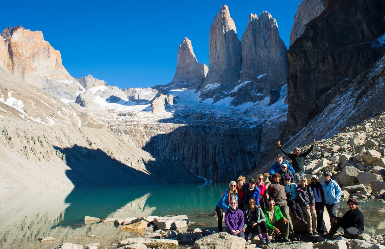 Everyone made it. The Torres don't care about sore legs. A large group of hikers posing on rocky moraine beside a turquoise glacial lake at the Base of the Towers, with the iconic granite spires of Torres del Paine rising sharply against a clear blue sky on the o trek patagonia circuit