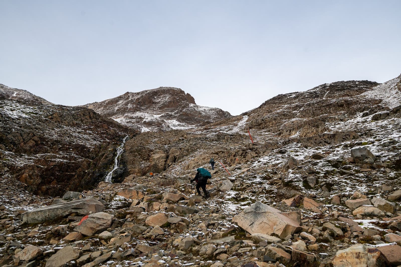 Rock by rock, the pass gets closer. This is what earned breakfast tastes like. Two trekkers with heavy packs pick their way up a snow-dusted boulder field on the o trek in patagonia, with a thin waterfall threading down the rocky gully and a jagged summit ridge rising into a pale grey sky above