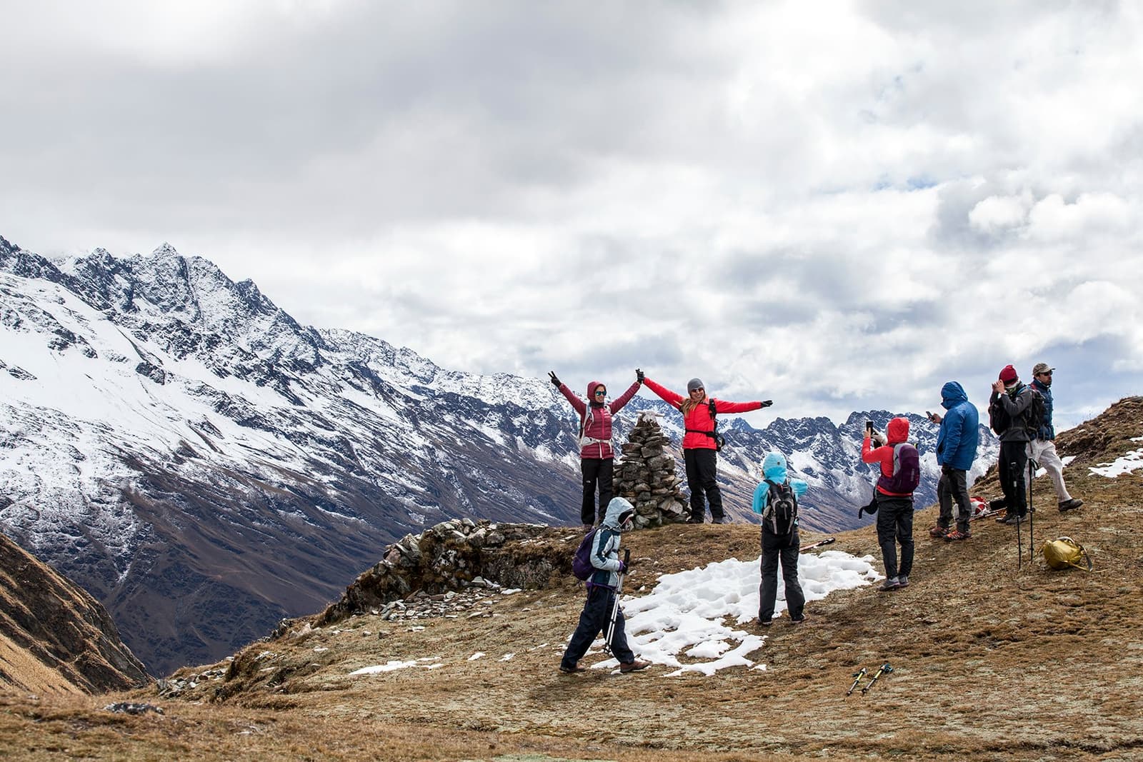 Arms up at the cairn. That earned it. A group of hikers celebrating at a stone cairn on the off the beaten path Salkantay trek, with snow-dusted peaks stretching across the horizon under an overcast sky