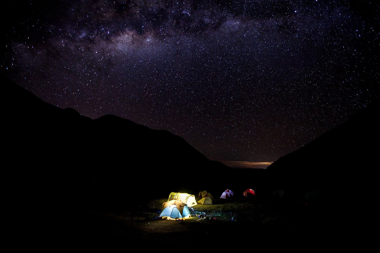 Your tent is the only light for miles, and the galaxy fills in the rest. Several illuminated tents glowing in the dark at a high-altitude campsite on the off-the-beaten-path Salkantay trek, with the full arc of the Milky Way stretching across the night sky above dark mountain silhouettes