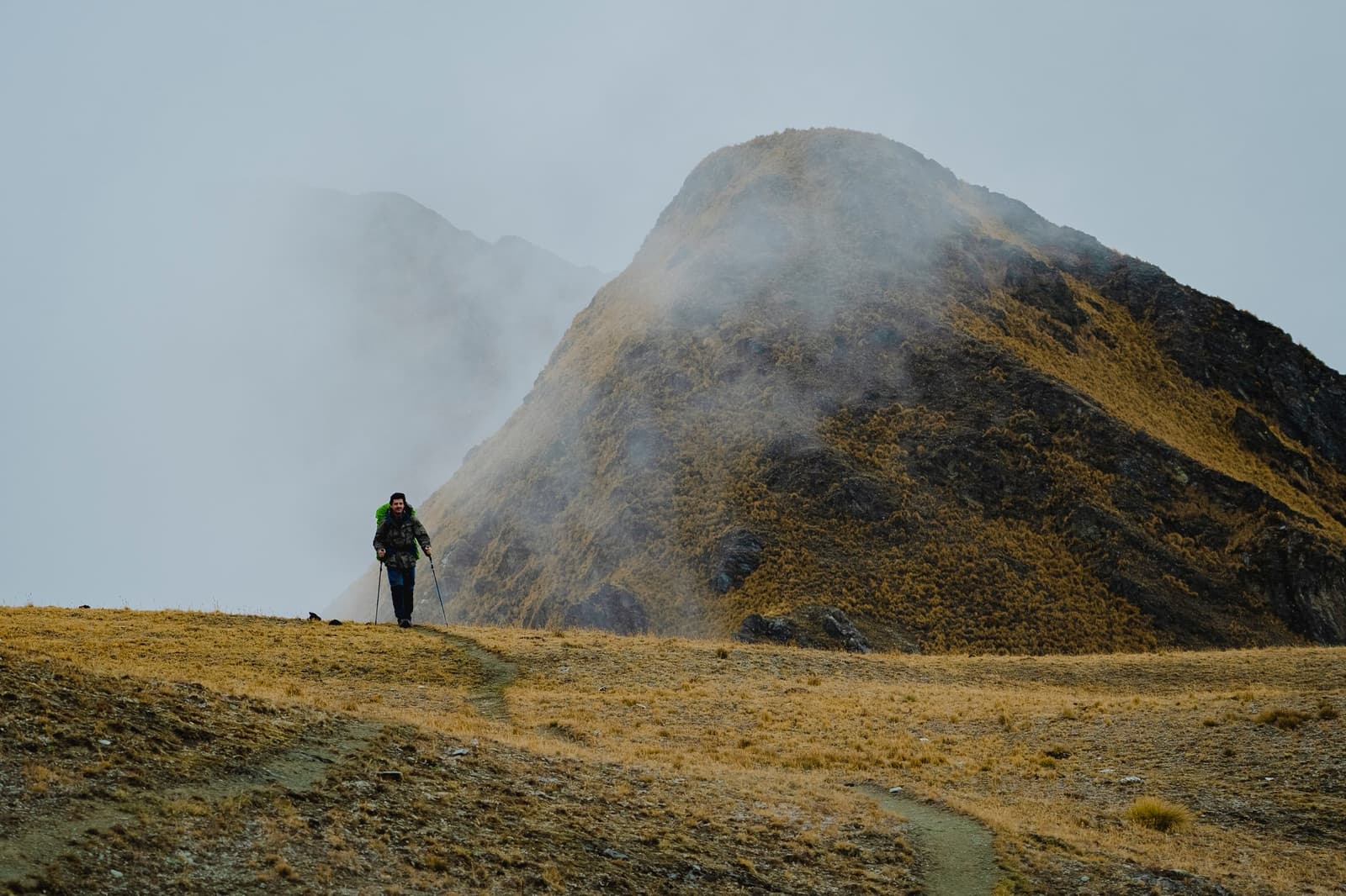 Clouds move fast up here, and so do smiles when no one else is on the trail. A solo hiker with trekking poles and a green backpack walks a narrow trail along a high-altitude grassland ridge on the off-the-beaten-path Salkantay trek, with mist rolling over steep rocky peaks behind them