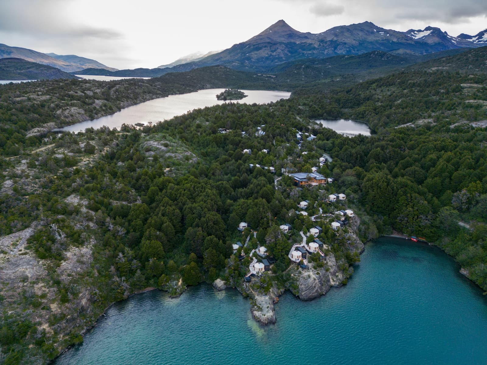 Tucked into the trees, but the lake and mountains are right there the whole time. Photo: Patagonia Camp Aerial view of Patagonia Camp's patagonia glamping yurts nestled in native lenga forest on a rocky peninsula above the turquoise waters of Lake Toro, with snow-capped Andean peaks in the background