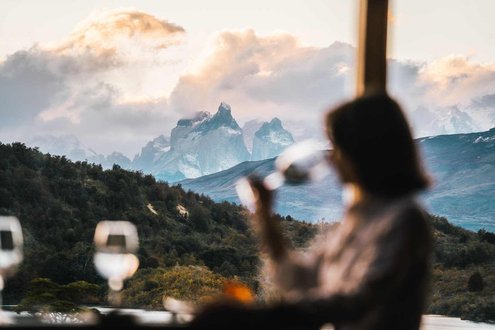 Good wine, better company, and that mountain just showing off in the background. Photo: Patagonia Camp A woman sips from a wine glass at the Patagonia Camp restaurant, with the snow-dusted granite peaks of the Paine Massif rising through clouds beyond the large window, wine glasses and a set table in the foreground