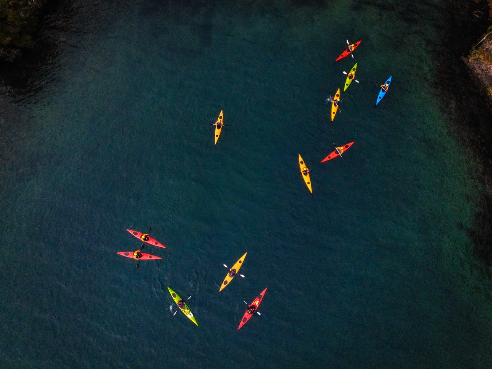 Eleven kayaks, eleven different ideas about the right pace. Photo: Patagonia Camp Aerial view of eleven colourful kayaks — red, yellow, green, and blue — scattered across the deep teal waters of Lake Toro during a guided patagonia camp morning paddle