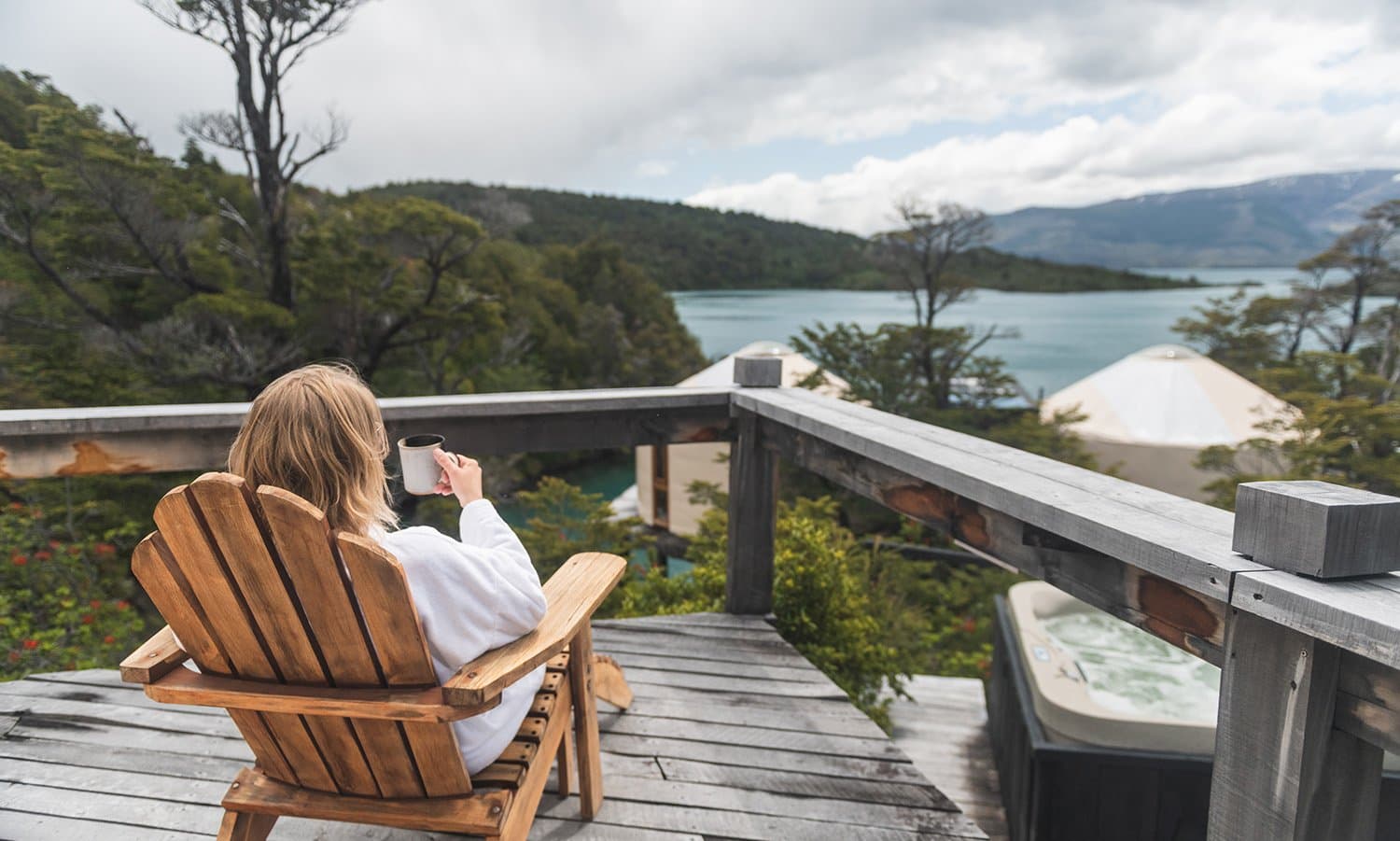 Coffee, a bathrobe, and nowhere you need to be for a while. Photo: Patagonia Camp A guest in a white bathrobe sits in a wooden Adirondack chair on a private deck at Patagonia Camp, holding a coffee mug and looking out over forested hillsides and a turquoise lake, with a private hot tub visible to the right
